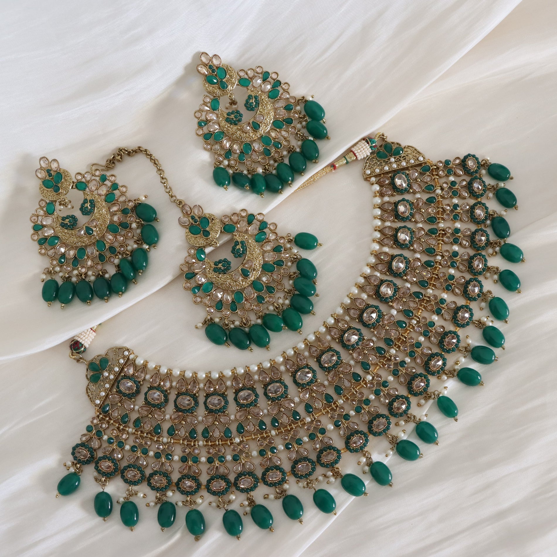 A traditional Indian jewelry set featuring a gold-plated necklace, matching earrings, and a maang tikka, embellished with green teardrop beads, clear crystals, and white faux pearls, displayed on a draped white fabric background.