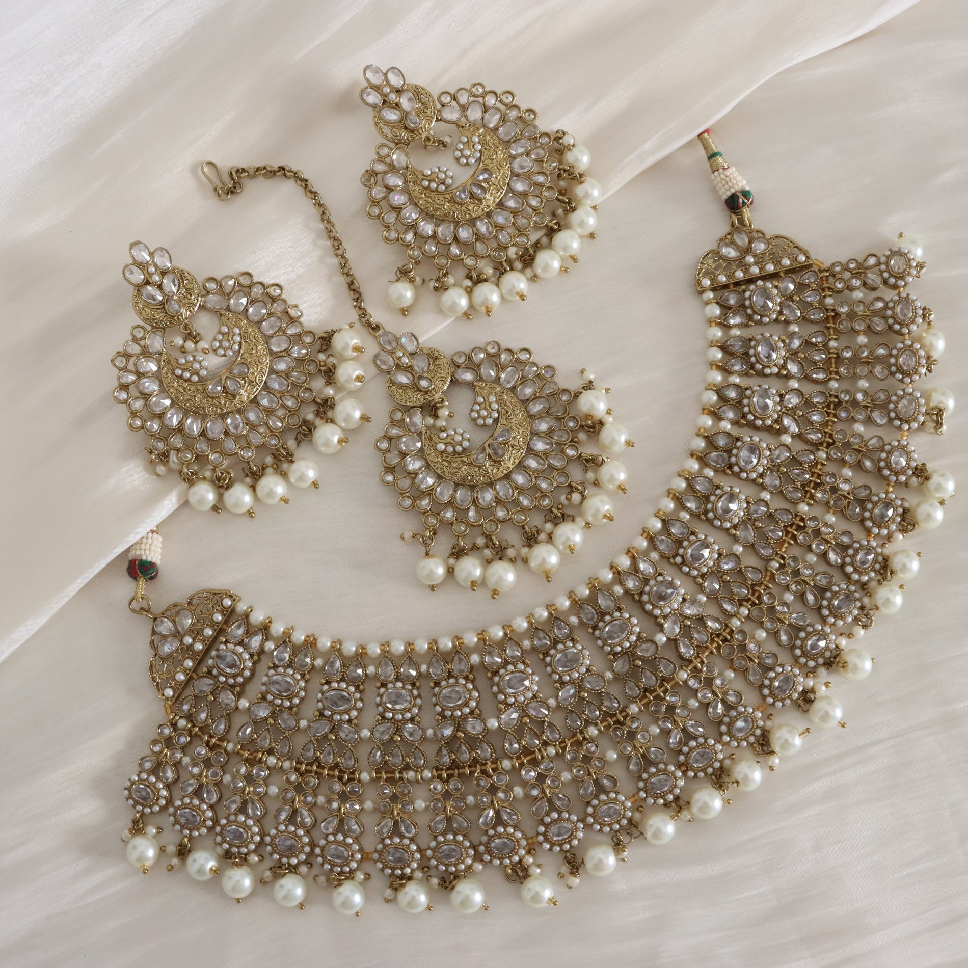 An ornate gold-toned traditional jewelry set with a crescent-shaped necklace, matching earrings, and a maang tikka, decorated with intricate beadwork, clear stones, and white faux pearls, arranged on a draped white fabric background.