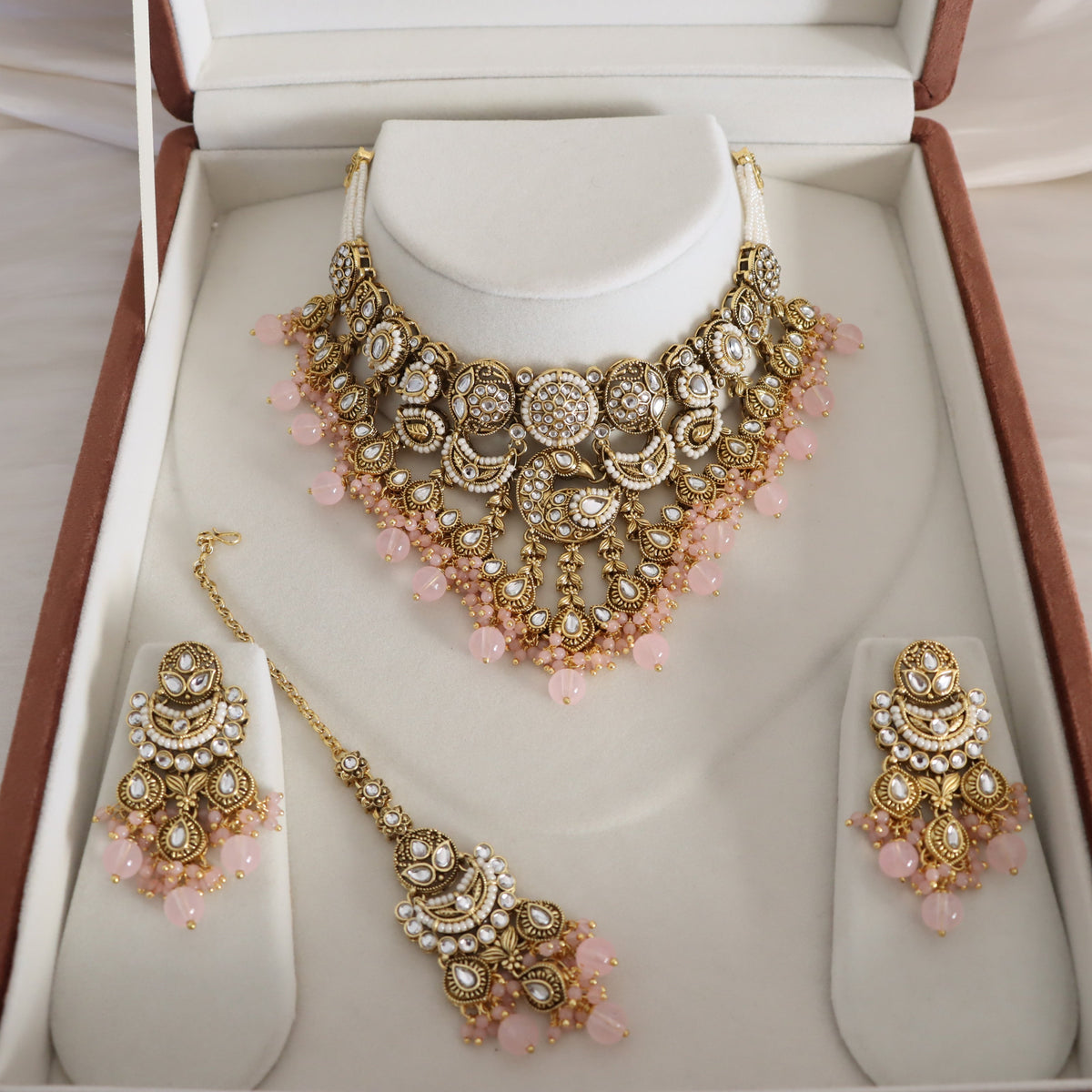 An ornate Indian ethnic jewelry set displayed inside an open presentation box. The set includes a gold-plated choker necklace with pink beads, matching drop earrings, and a maang tikka, all decorated with white faux pearls and clear stones.