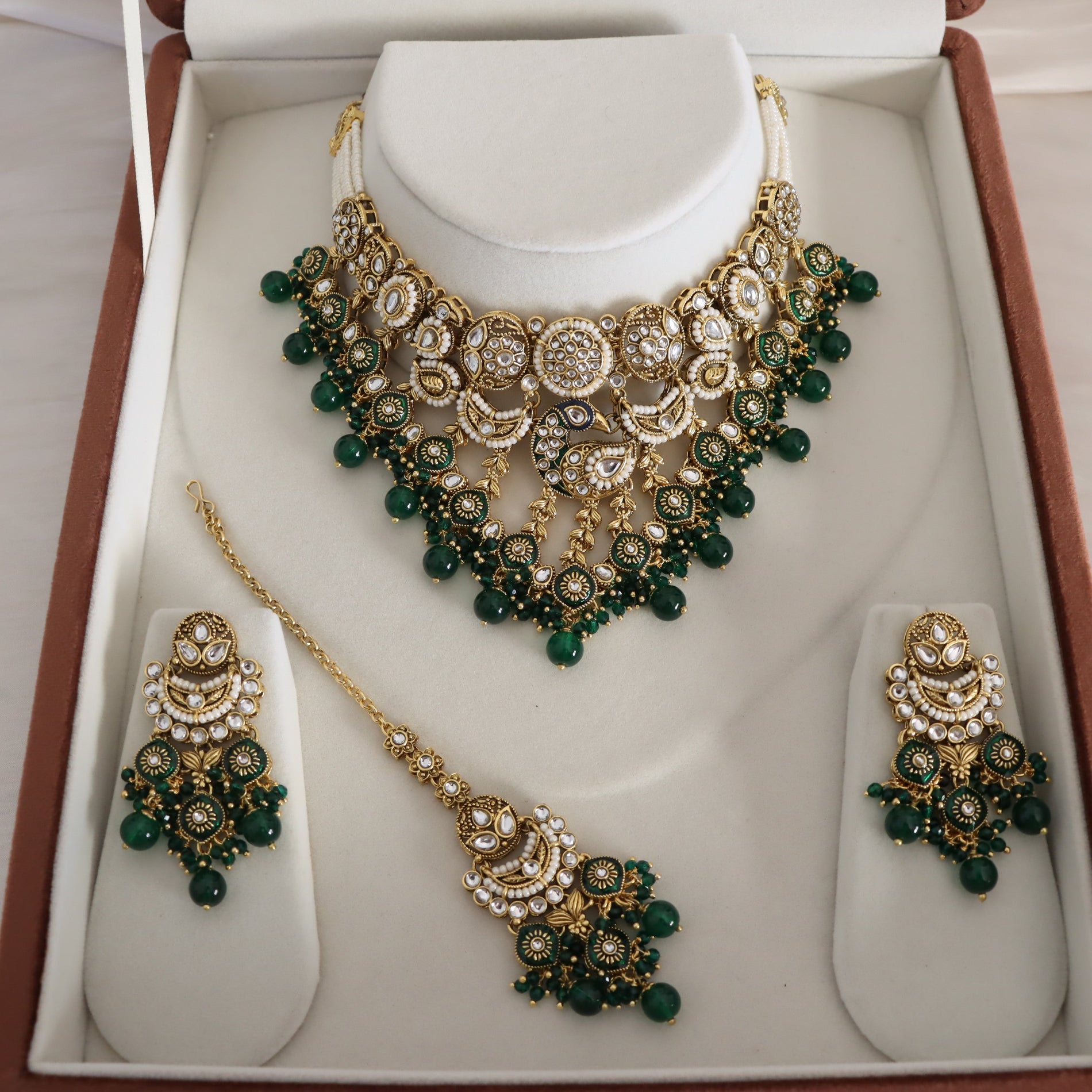 Gold and green traditional Indian jewelry set displayed in a white jewelry box, including a statement necklace, matching earrings, and a maang tikka, with clear stones and green beads.