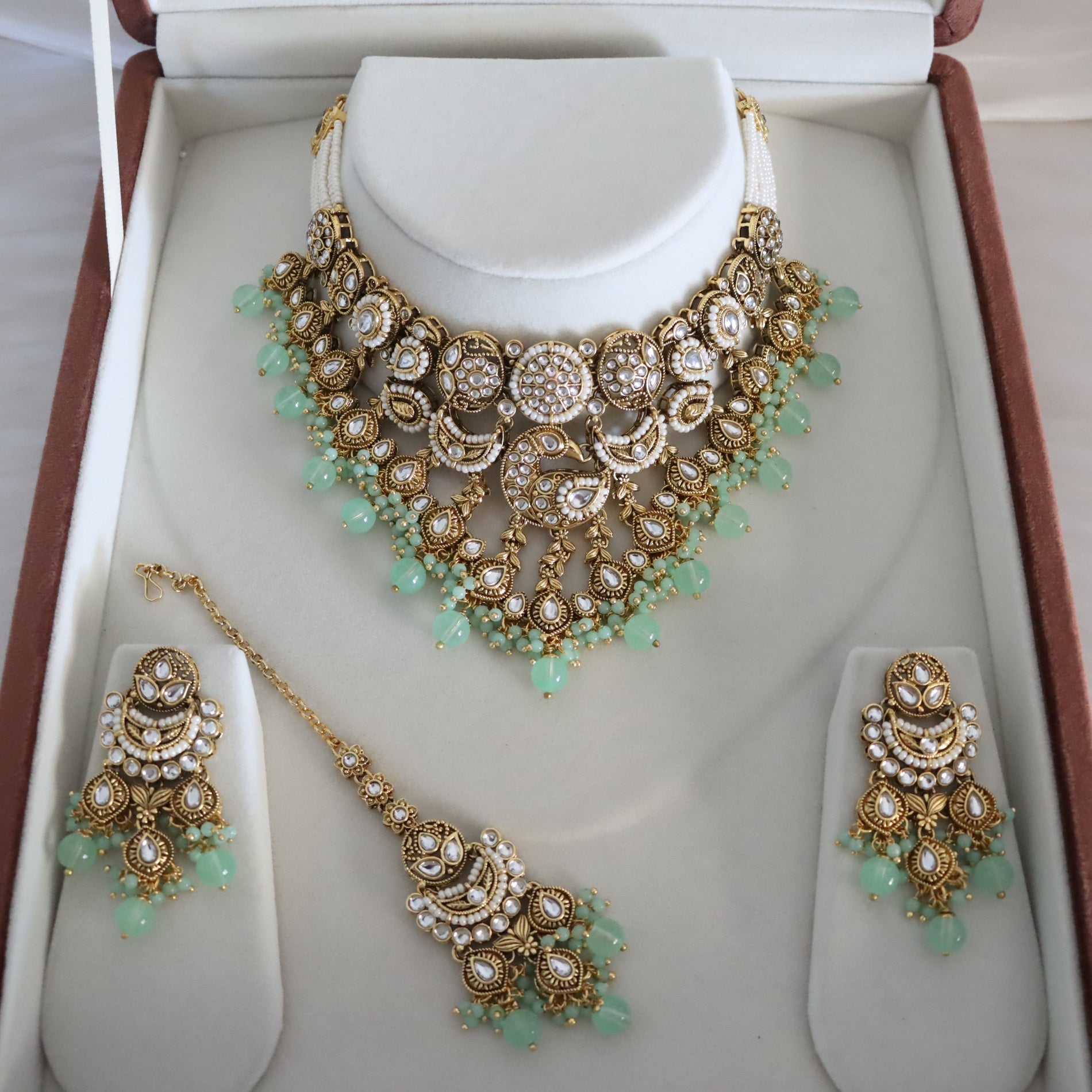 An ornate gold-toned jewelry set displayed inside an open presentation box, including a statement necklace on a bust, matching earrings, and a dangling maang tikka, decorated with white stones and light green bead accents.