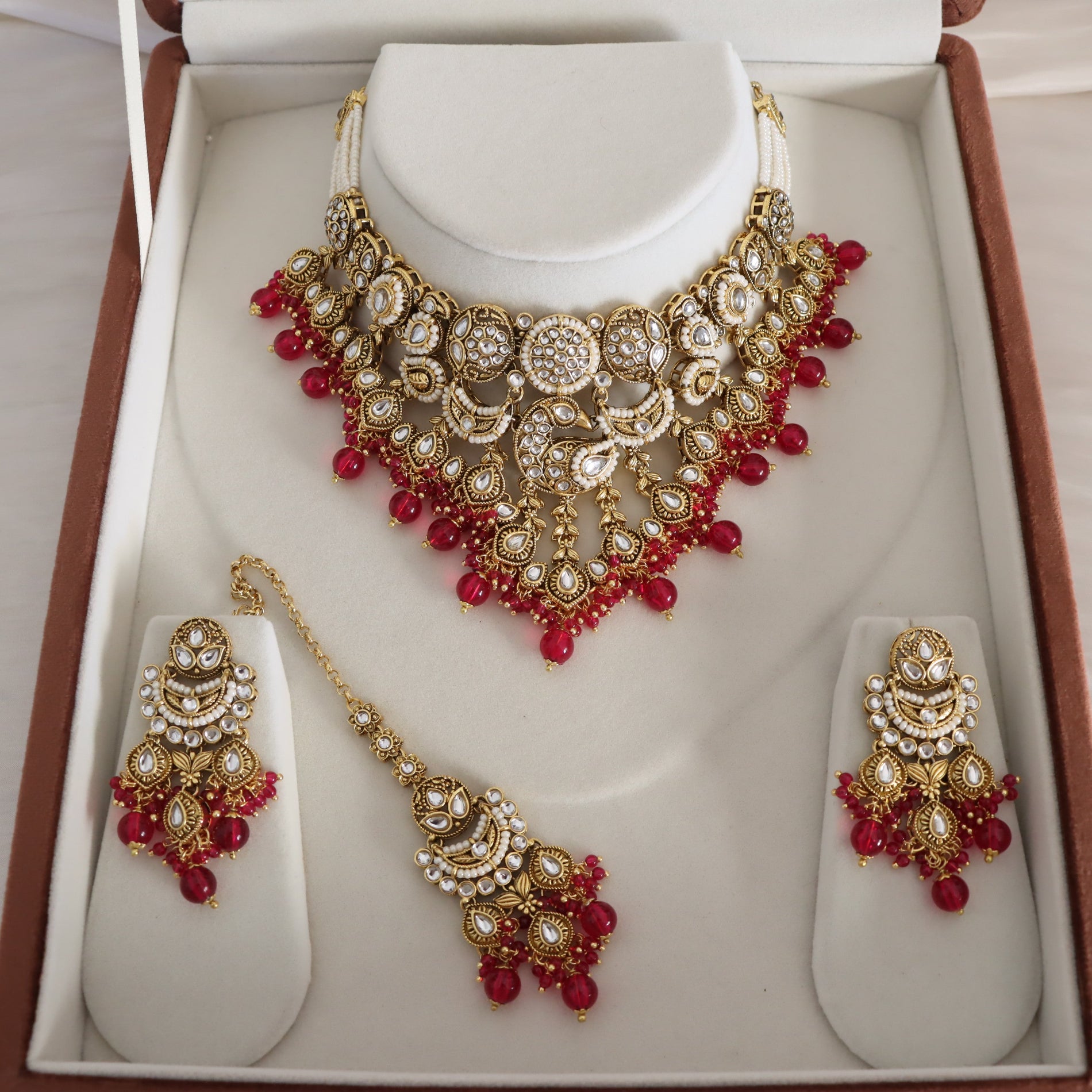 An ornate Indian traditional jewelry set displayed inside its brown presentation box. The set includes a gold-plated choker necklace with red beads and white stones, matching drop earrings, and a maang tikka, all arranged on white display stands.