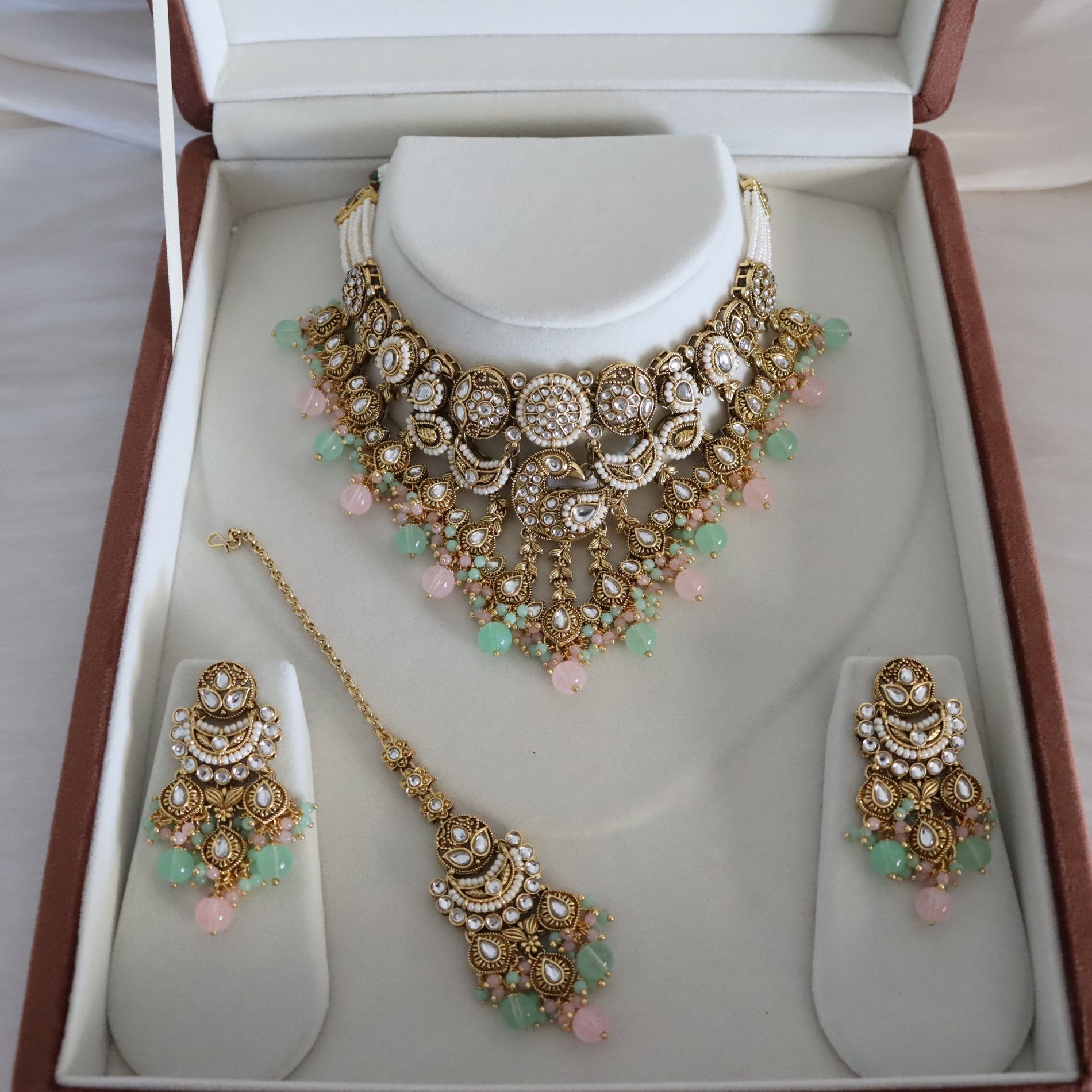 Gold-toned Indian ethnic jewelry set including a choker necklace, matching pendant, and earrings, displayed on white stand inserts inside an open presentation box.