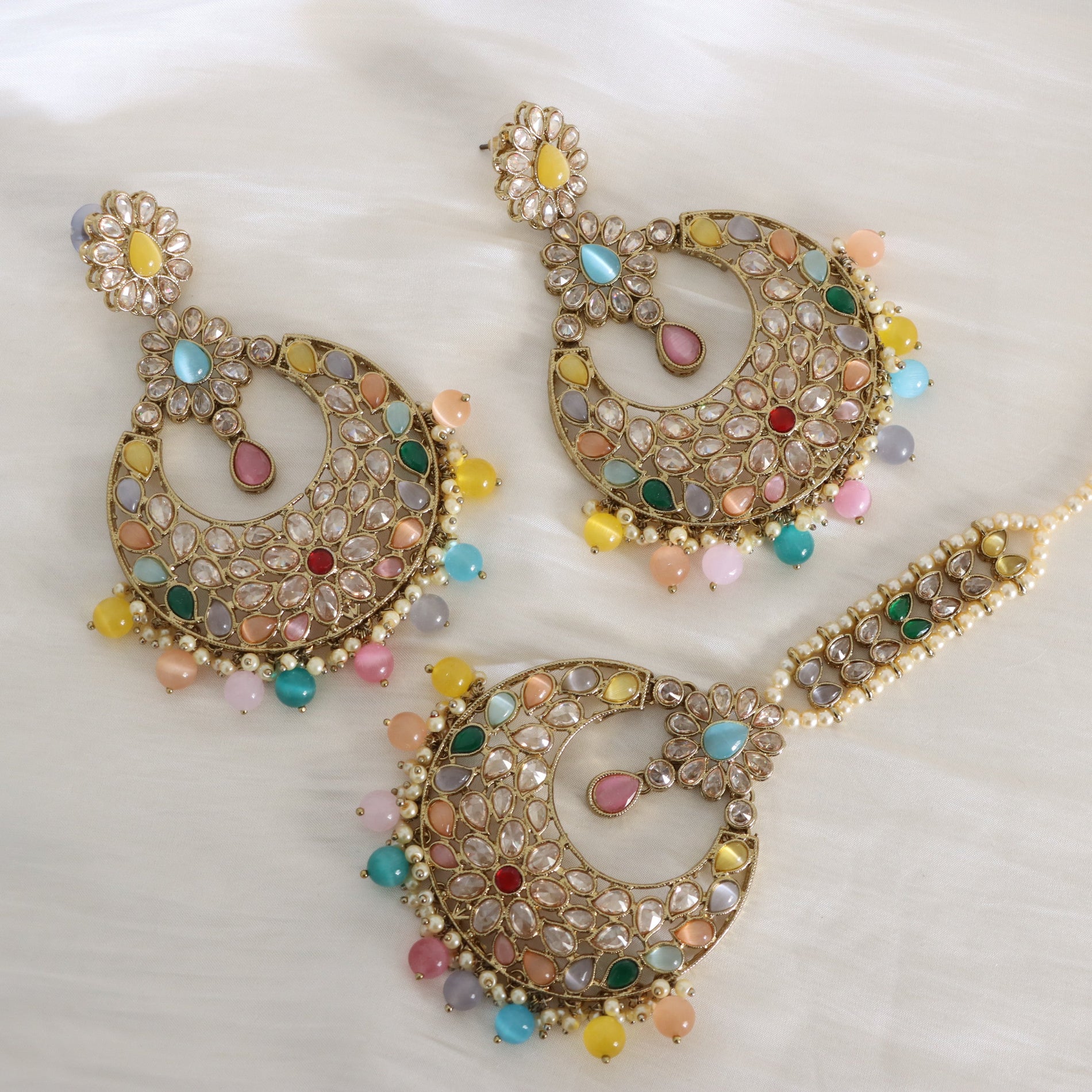 Gold-toned traditional Indian jhumka earrings with multicolored stone embellishments, displayed on a soft white fabric background.