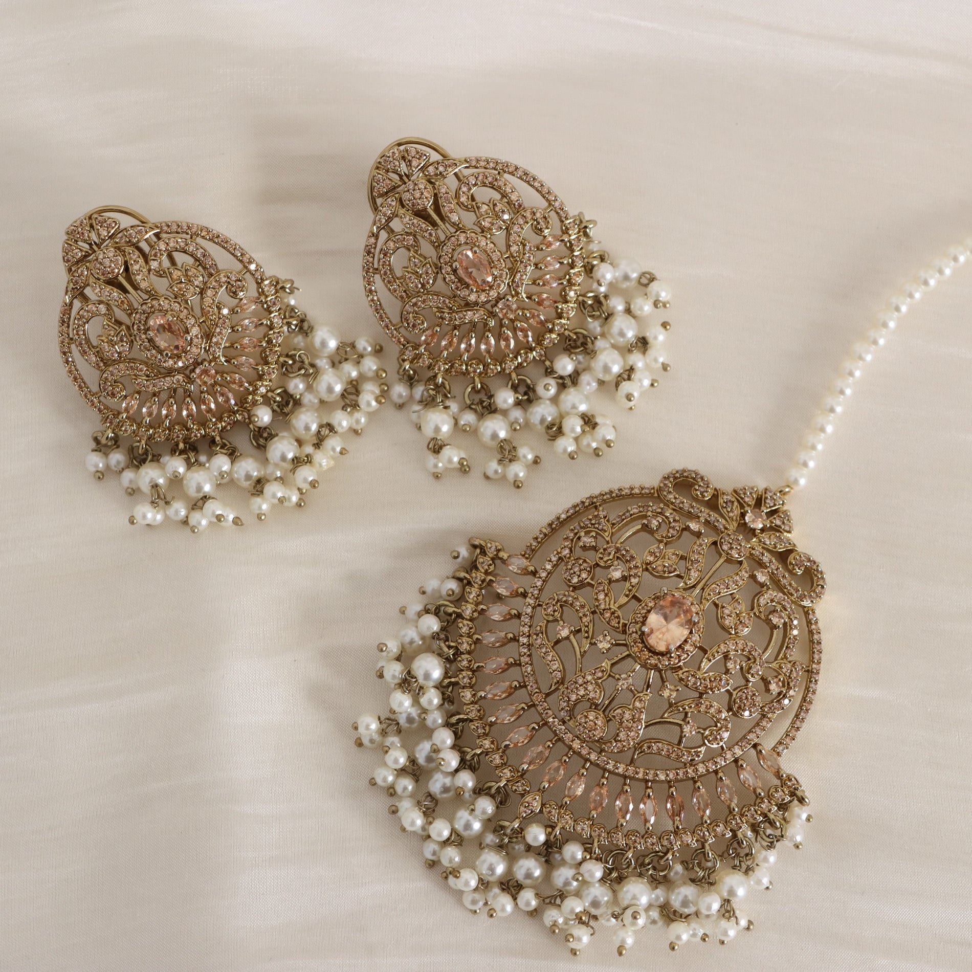 An ornate gold-toned traditional jewelry set featuring a large circular pendant, a matching earring, and a pearl-beaded headpiece, arranged on a soft cream fabric background.