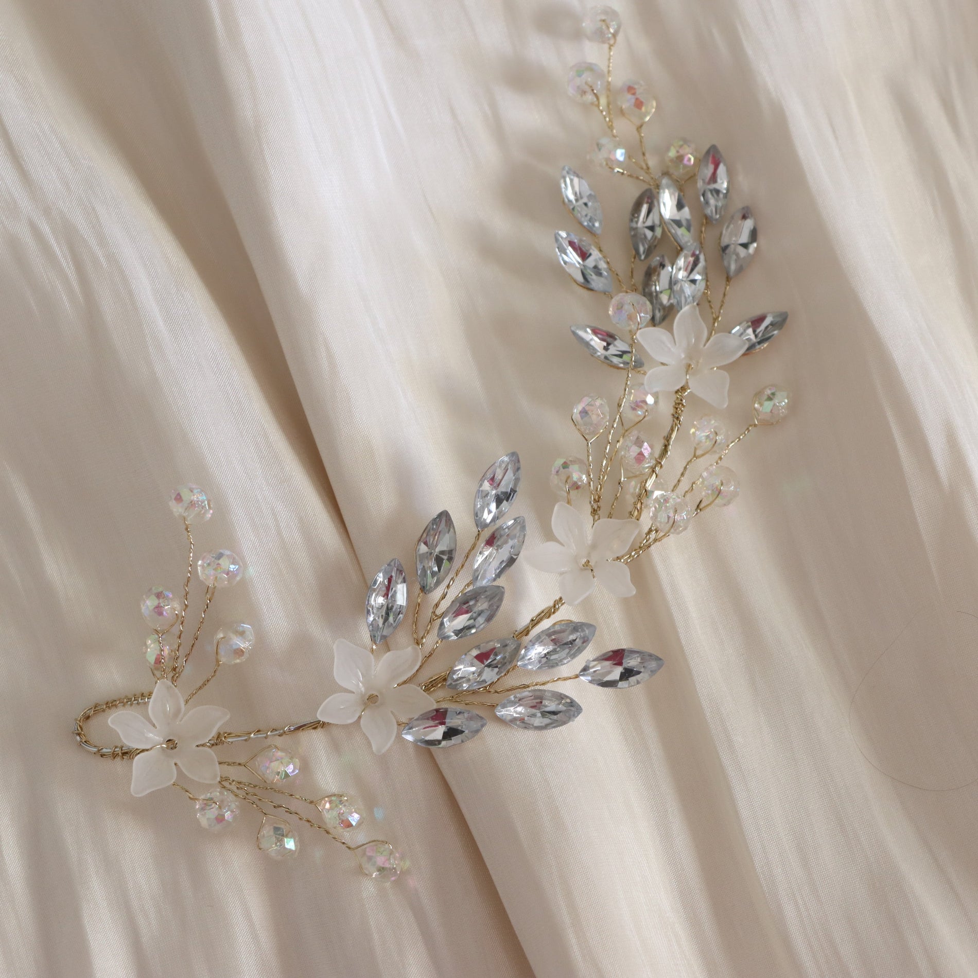 A pair of bridal hairpieces with gold-tone wire branches, clear rhinestones, and white faux flowers, displayed on draped champagne-colored fabric.