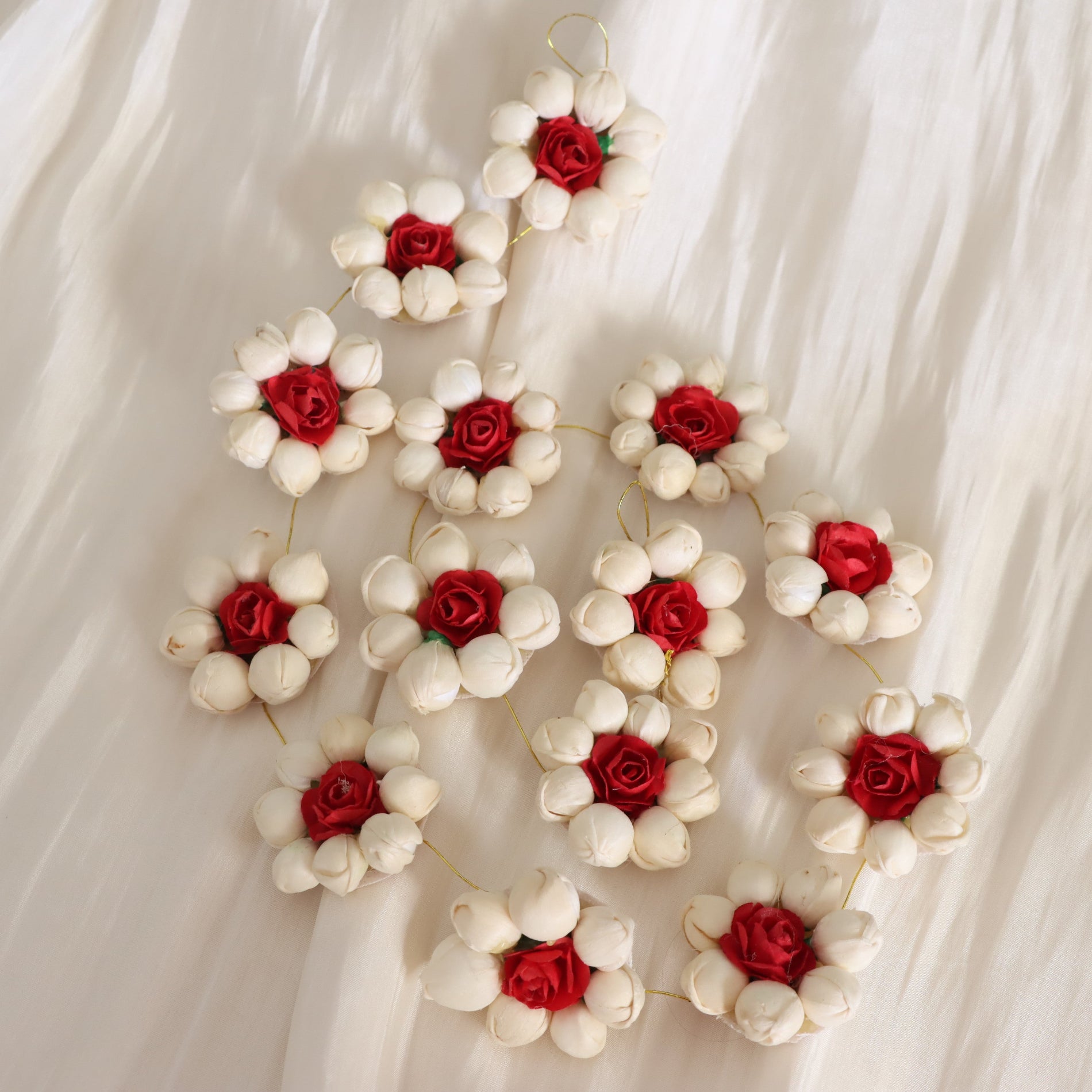 A decorative garland made of multiple small white flower blossoms with red rose centers, arranged on a soft white fabric background.