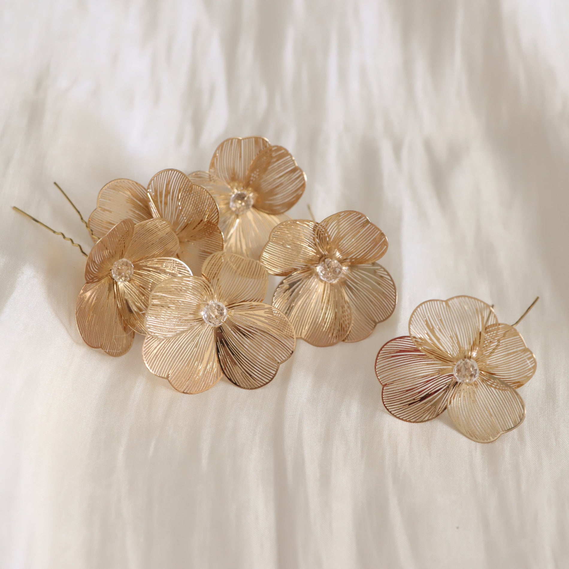 A set of gold-tone flower hair pins with delicate wire detailing and clear crystal centers, arranged on a soft white textured fabric background.