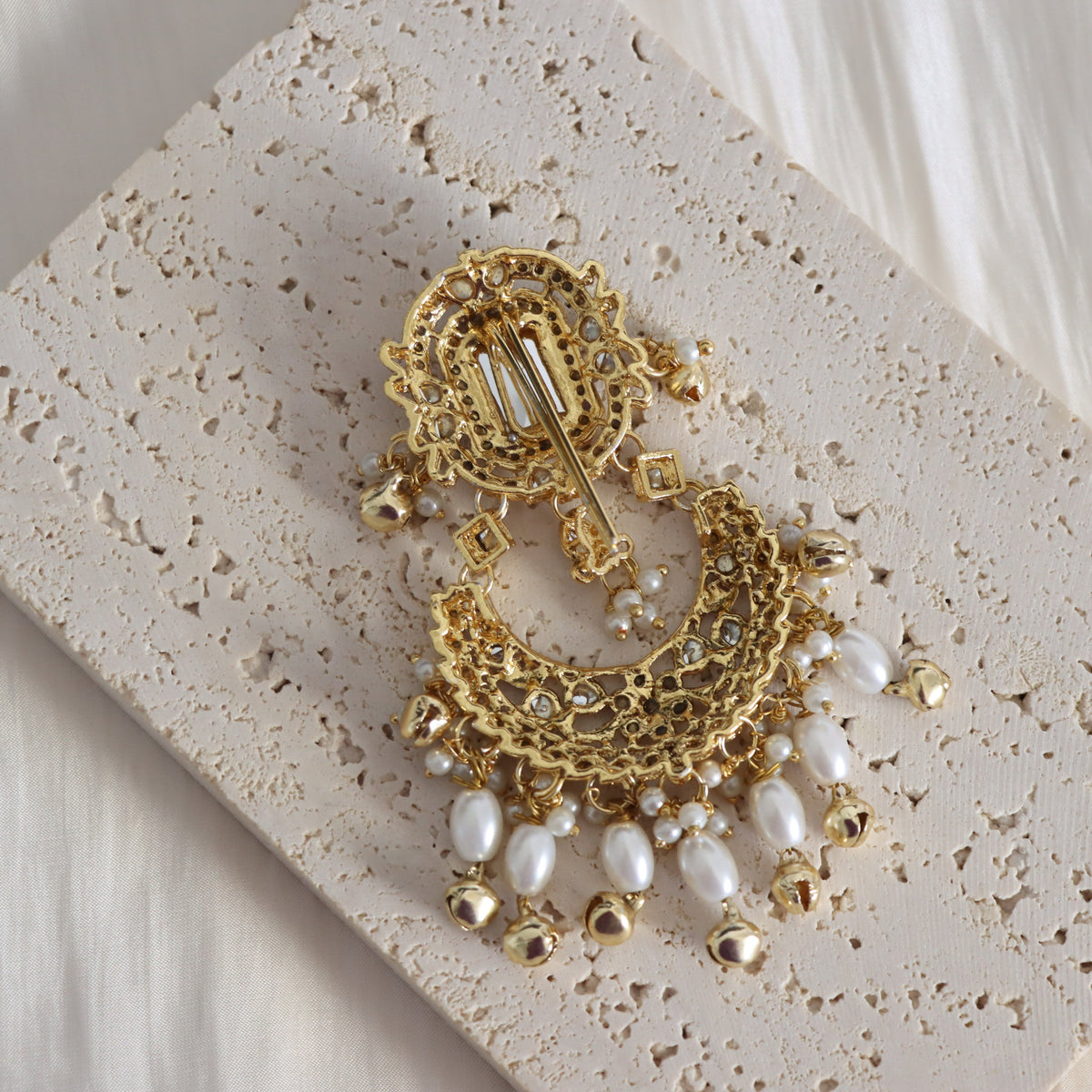 An ornate gold-toned statement earring with intricate filigree detailing, small clear rhinestones, and dangling white faux pearls, displayed on a textured beige stone slab.