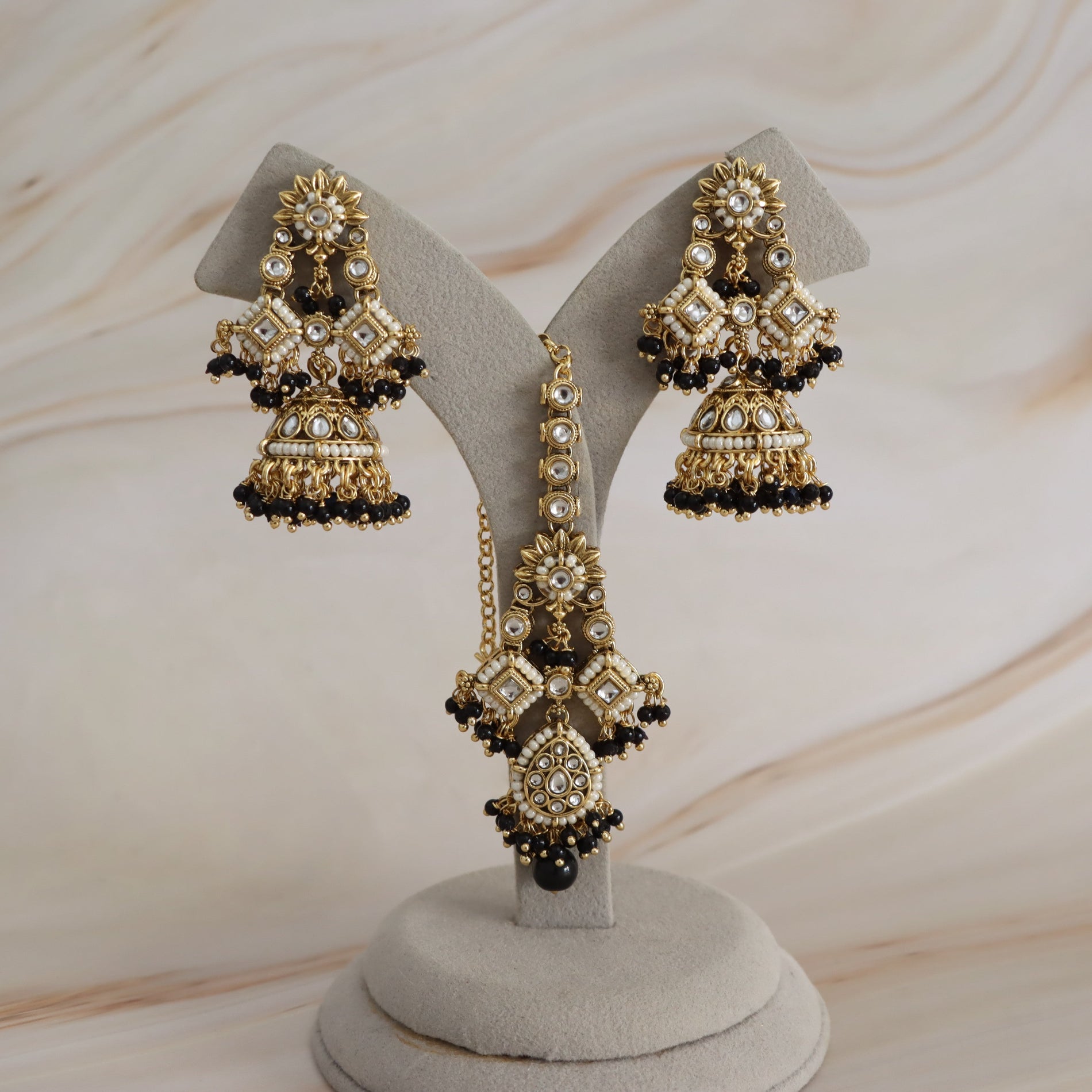 Gold-plated Indian jhumka earrings with black beads and white faux pearls, displayed on a gray velvet jewelry stand against a light beige marble background.
