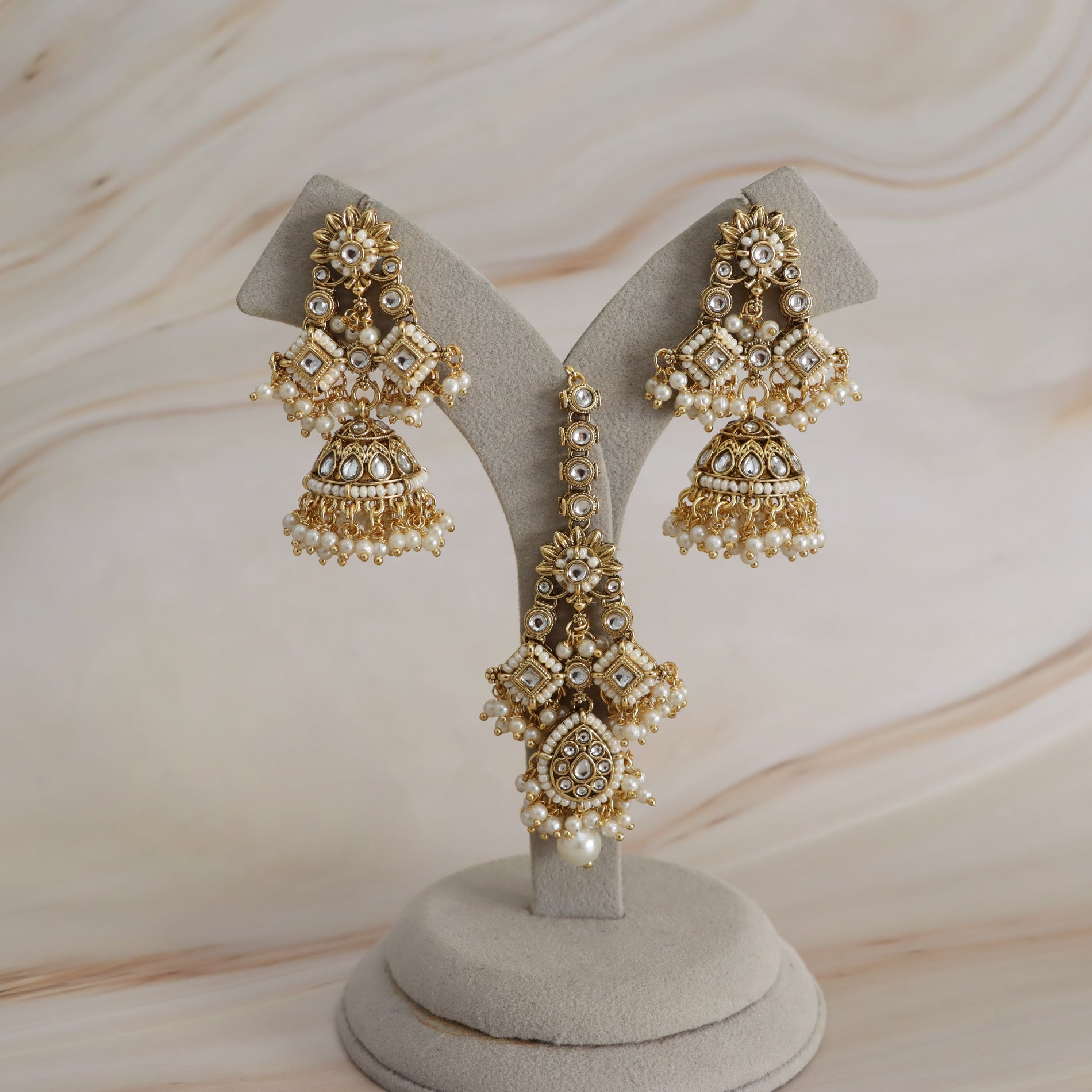 A pair of ornate gold-toned jhumka earrings with white faux pearls and clear rhinestone embellishments, displayed on a light gray velvet jewelry stand against a soft beige marble background.