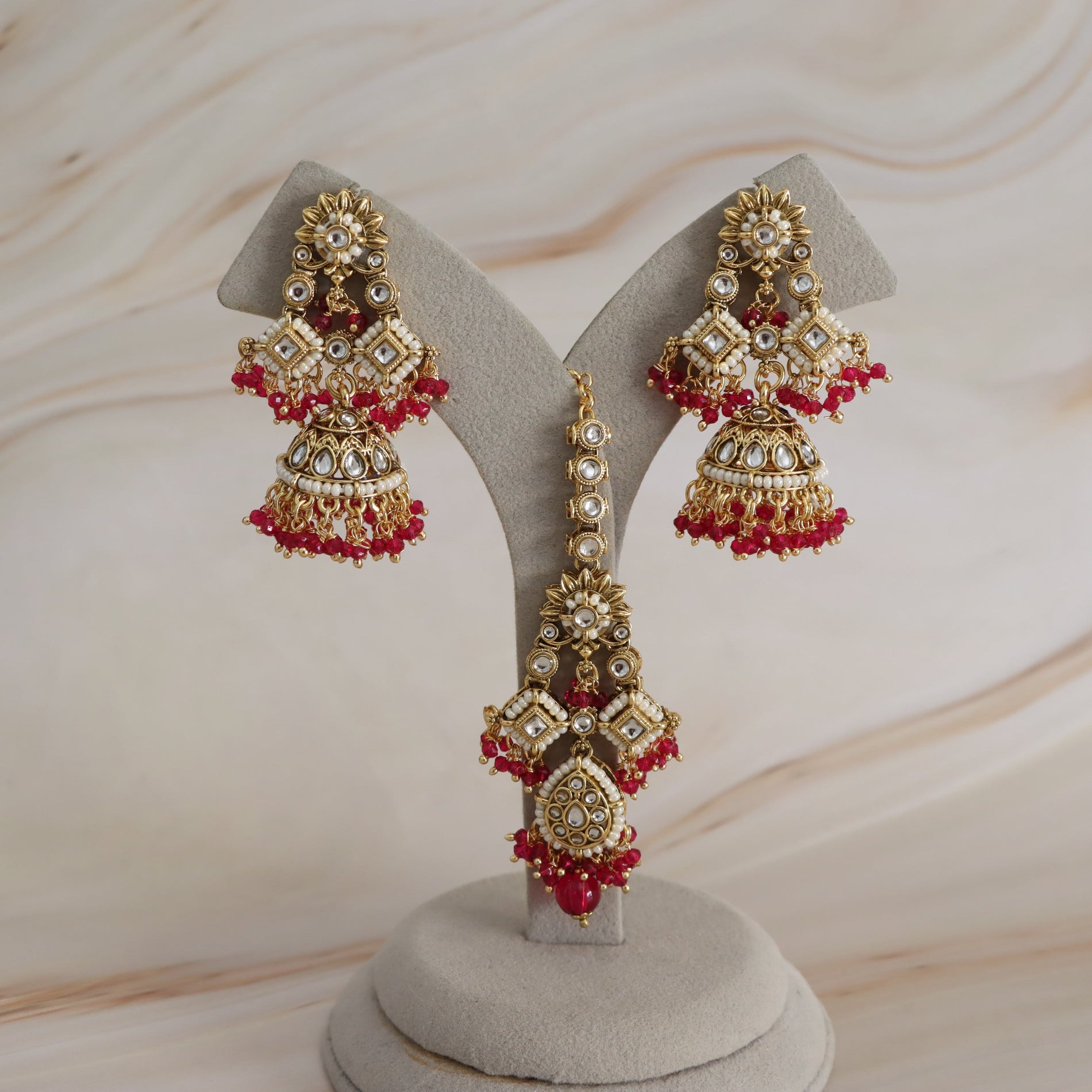 Gold-plated Indian jhumka earrings with red bead borders and clear stone embellishments, displayed on a gray velvet jewelry stand against a light wood background.
