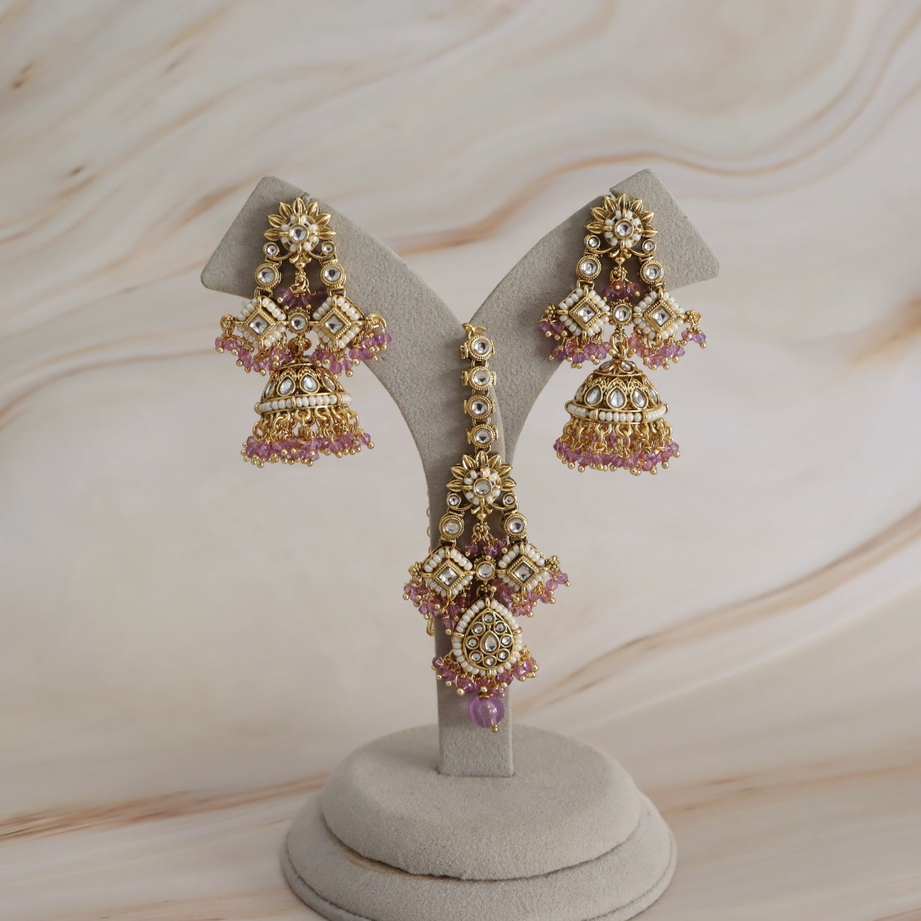 A pair of ornate gold-toned jhumka earrings with pink stone accents and clear crystal details, displayed on a beige jewelry stand against a light marble-patterned background.