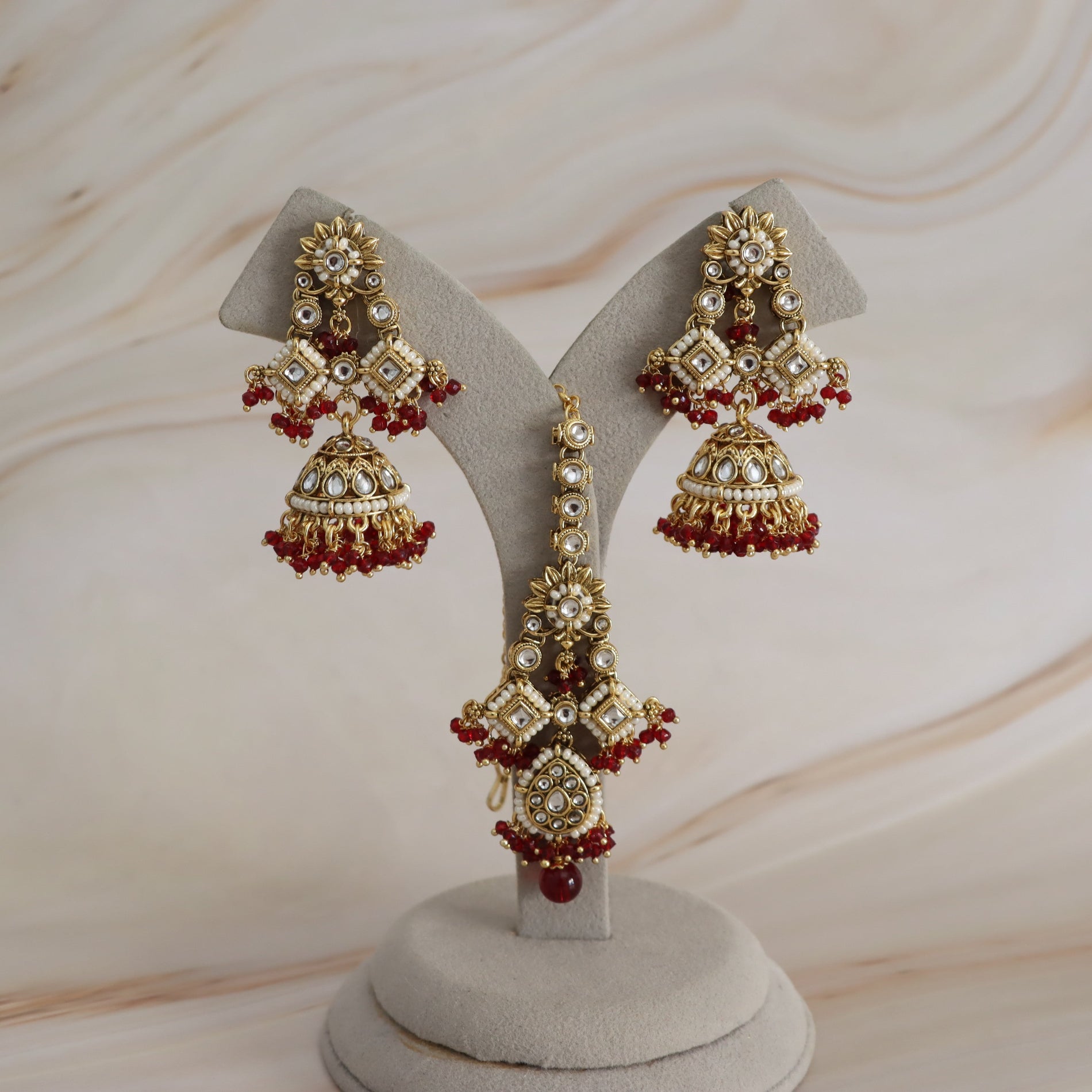 Gold-toned traditional Indian jhumka earrings with red bead borders and white stone accents, displayed on a beige jewelry stand against a soft beige marble background.