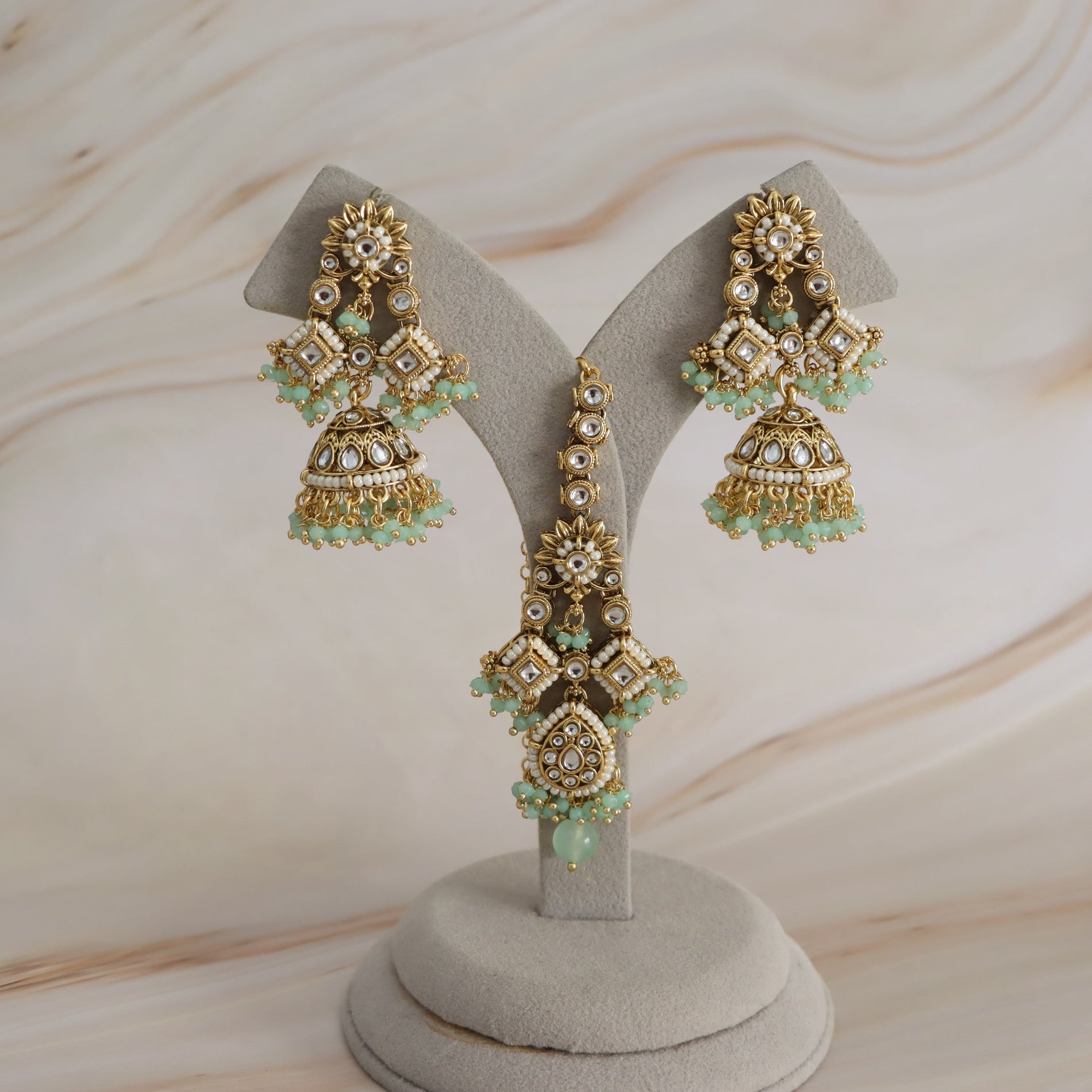 Gold-toned traditional Indian jhumka earrings with green bead accents and clear stones, displayed on a gray velvet jewelry stand against a light beige marble background.