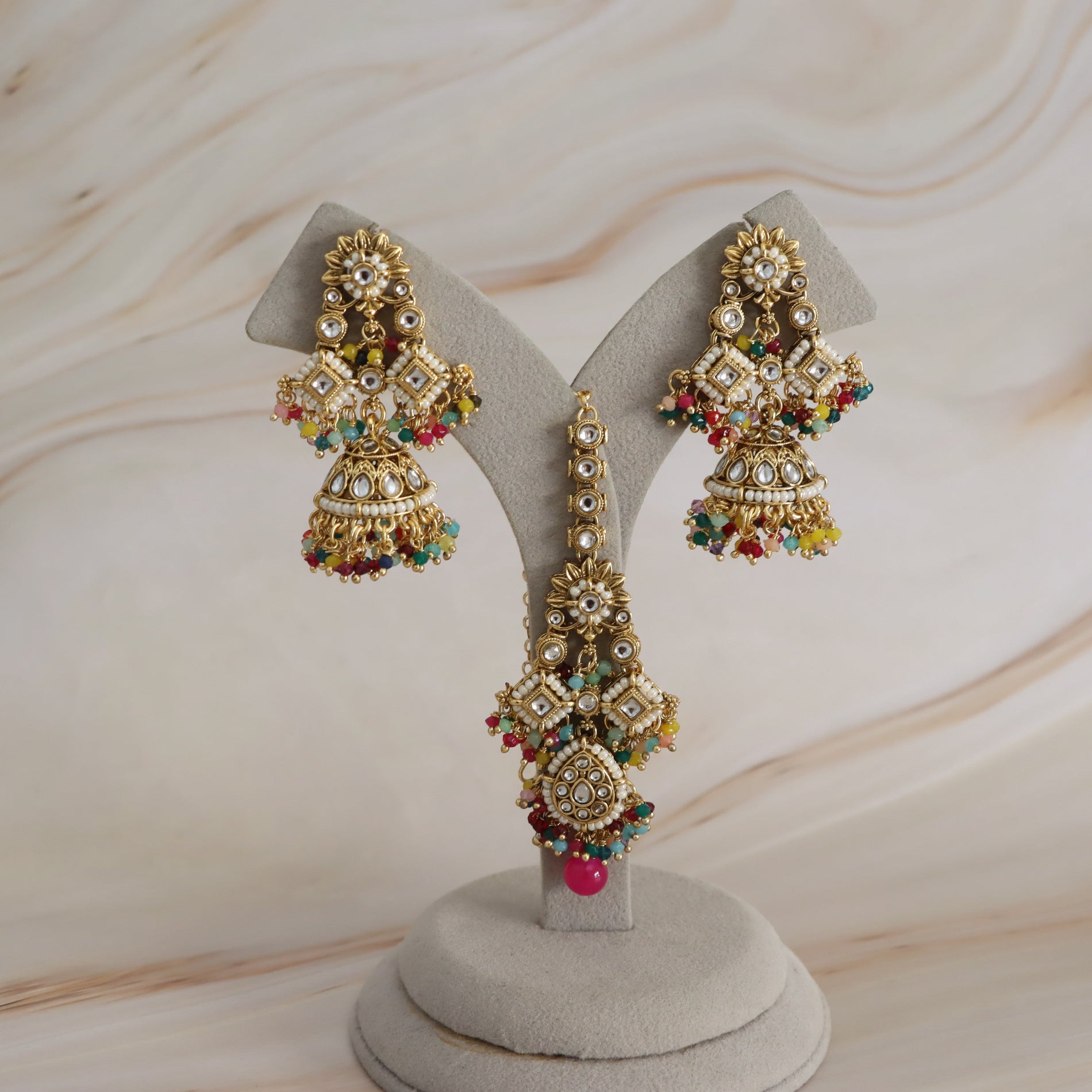 Gold-toned traditional Indian jhumka earrings with intricate stone embellishments and colorful bead accents, displayed on a gray jewelry stand against a light marble-patterned background.