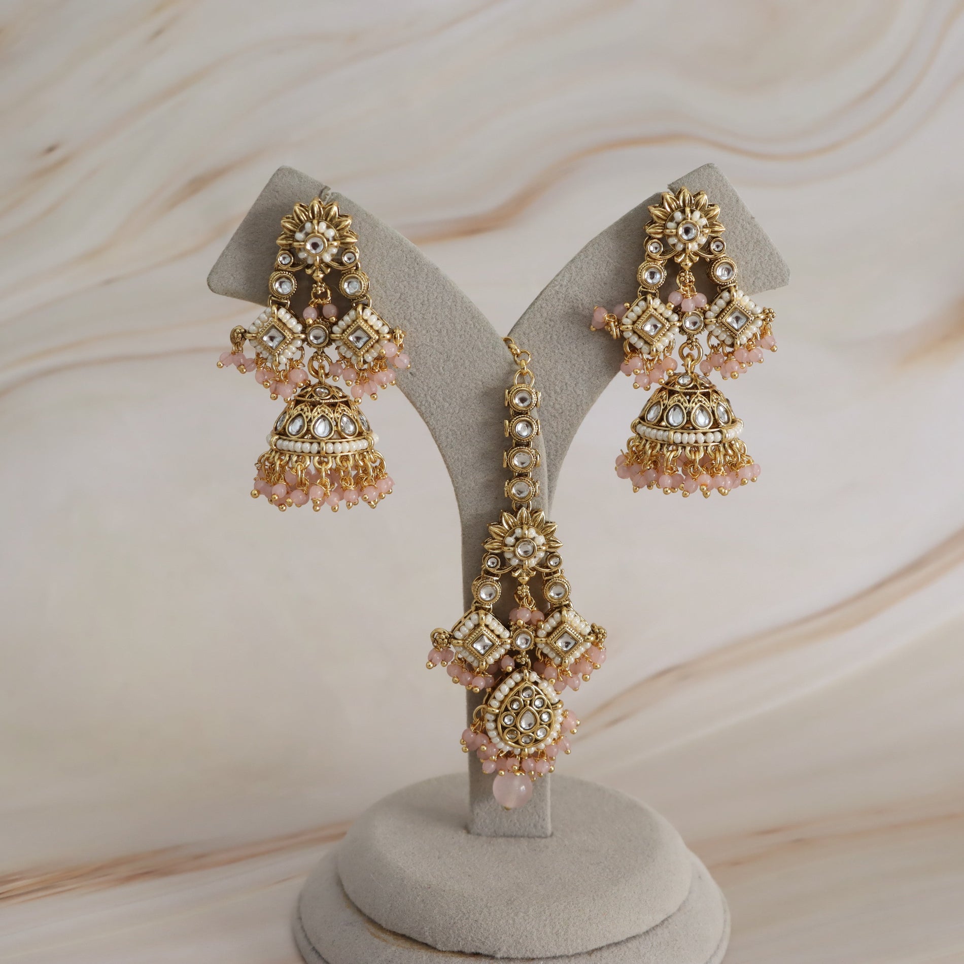 Gold-toned traditional Indian jhumka earrings with pink bead embellishments and clear stones, displayed on a gray velvet jewelry stand against a light beige marble background.