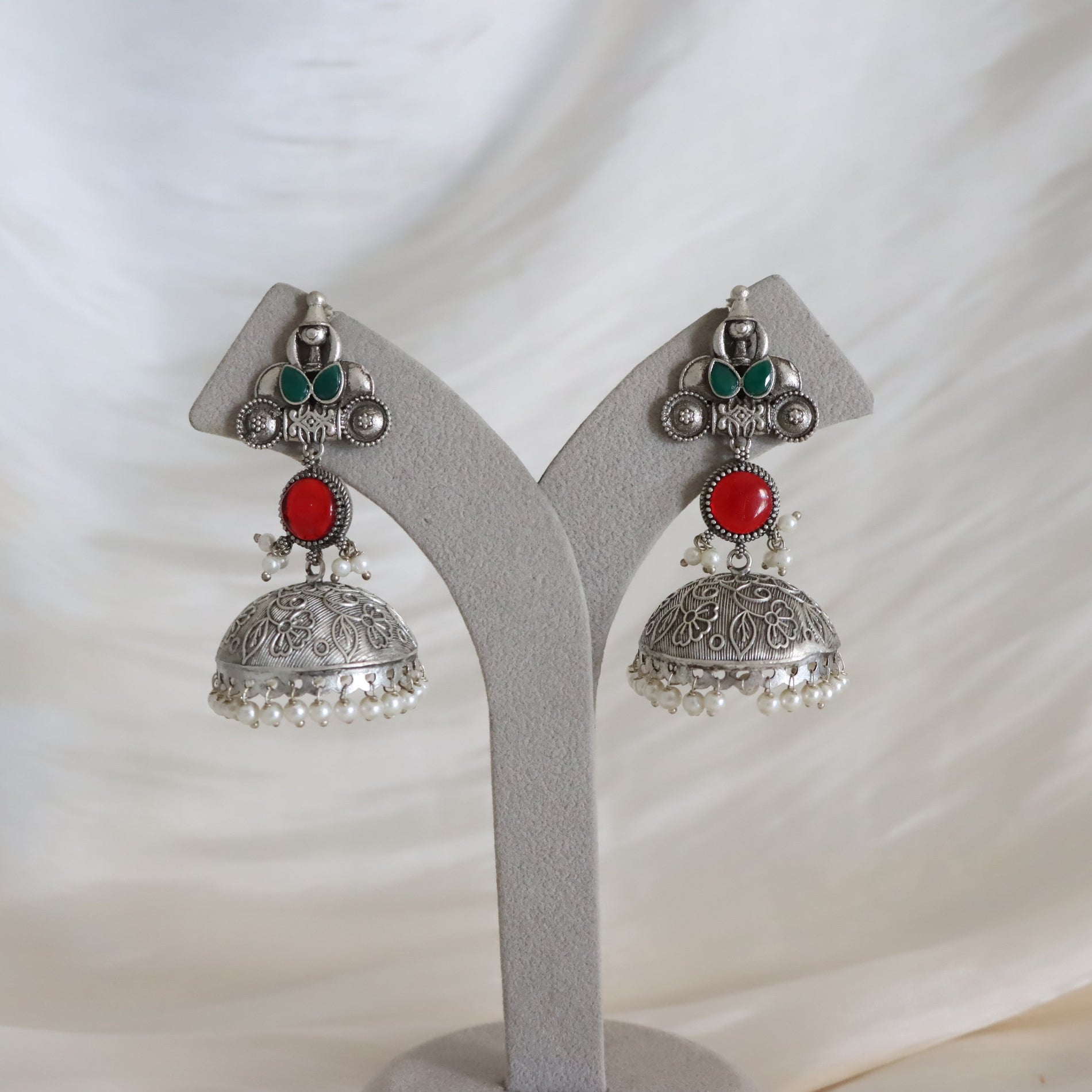 A pair of silver-toned Indian jhumka earrings with red and green stone accents, displayed on a gray Y-shaped jewelry stand against a draped white fabric background.