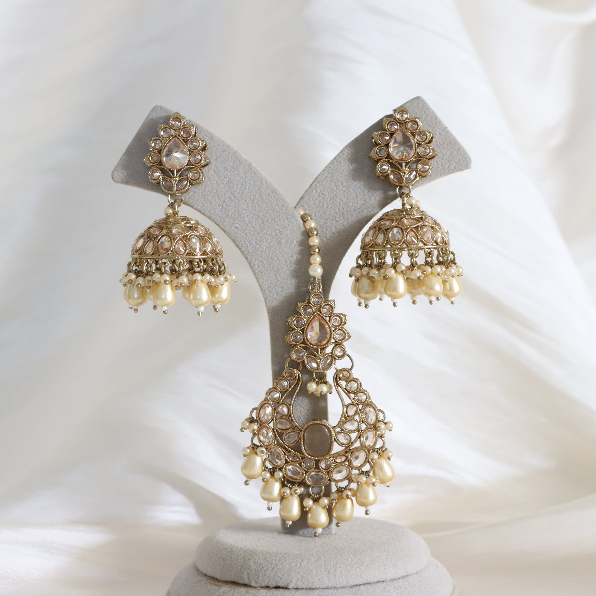 Gold-toned traditional Indian jhumka earrings with cream pearl-like beads and clear stones, displayed on a light gray jewelry stand against a soft white fabric background.