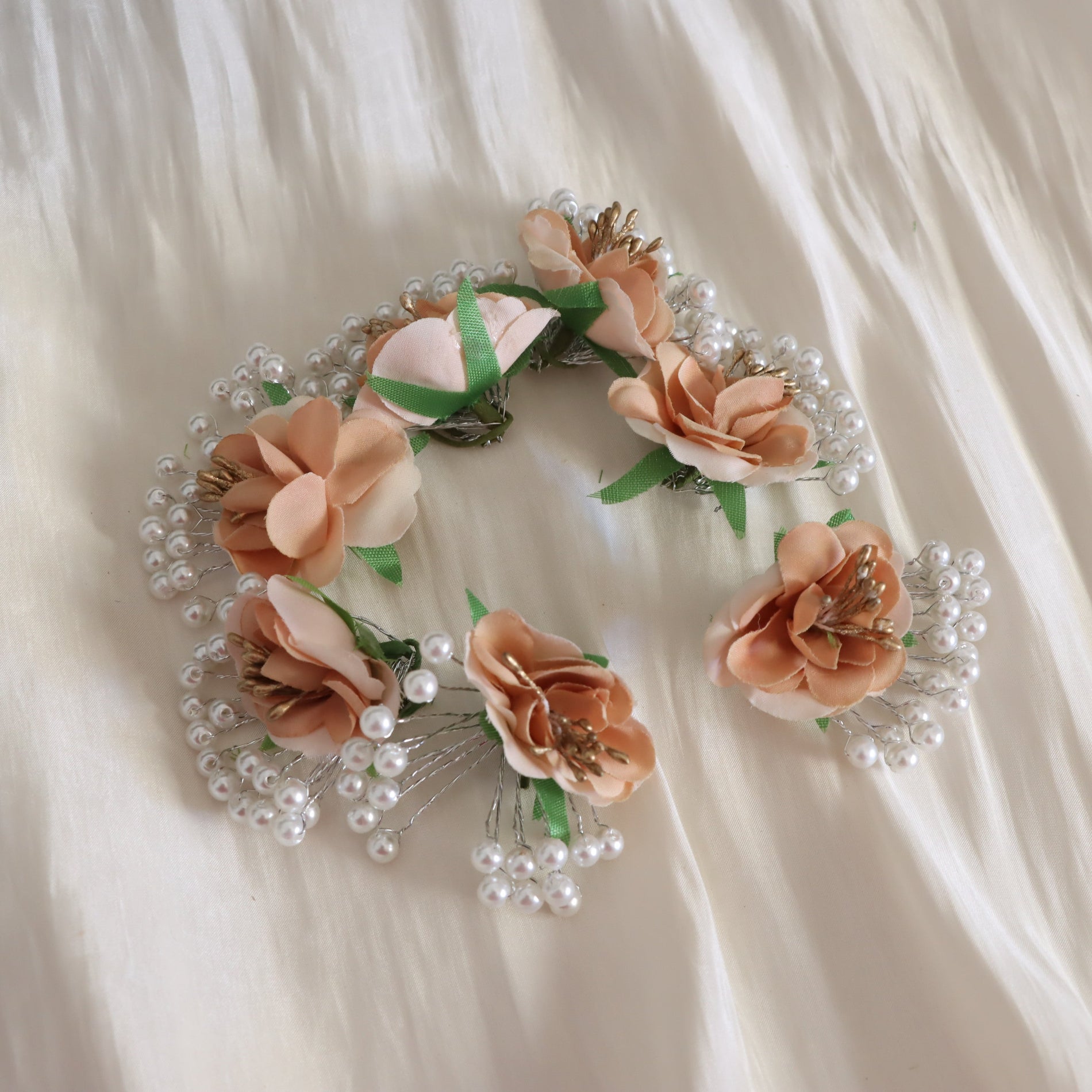A decorative floral headpiece or wreath made of peach and blush fabric flowers, green ribbon accents, and clusters of white faux pearls, arranged on a soft cream fabric background.