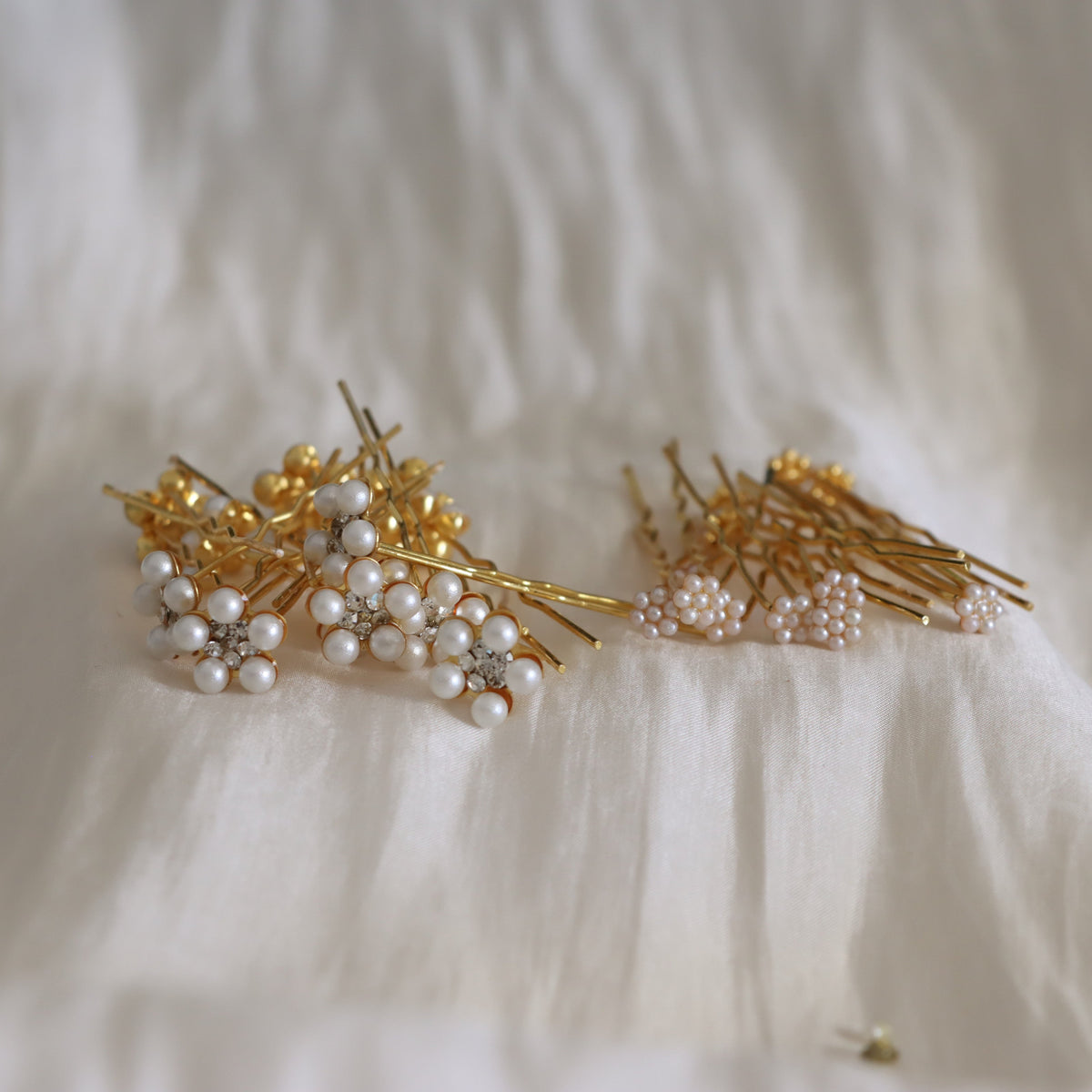 A set of gold hair pins decorated with clusters of white faux pearls and small clear rhinestones, arranged on a soft white textured fabric background.