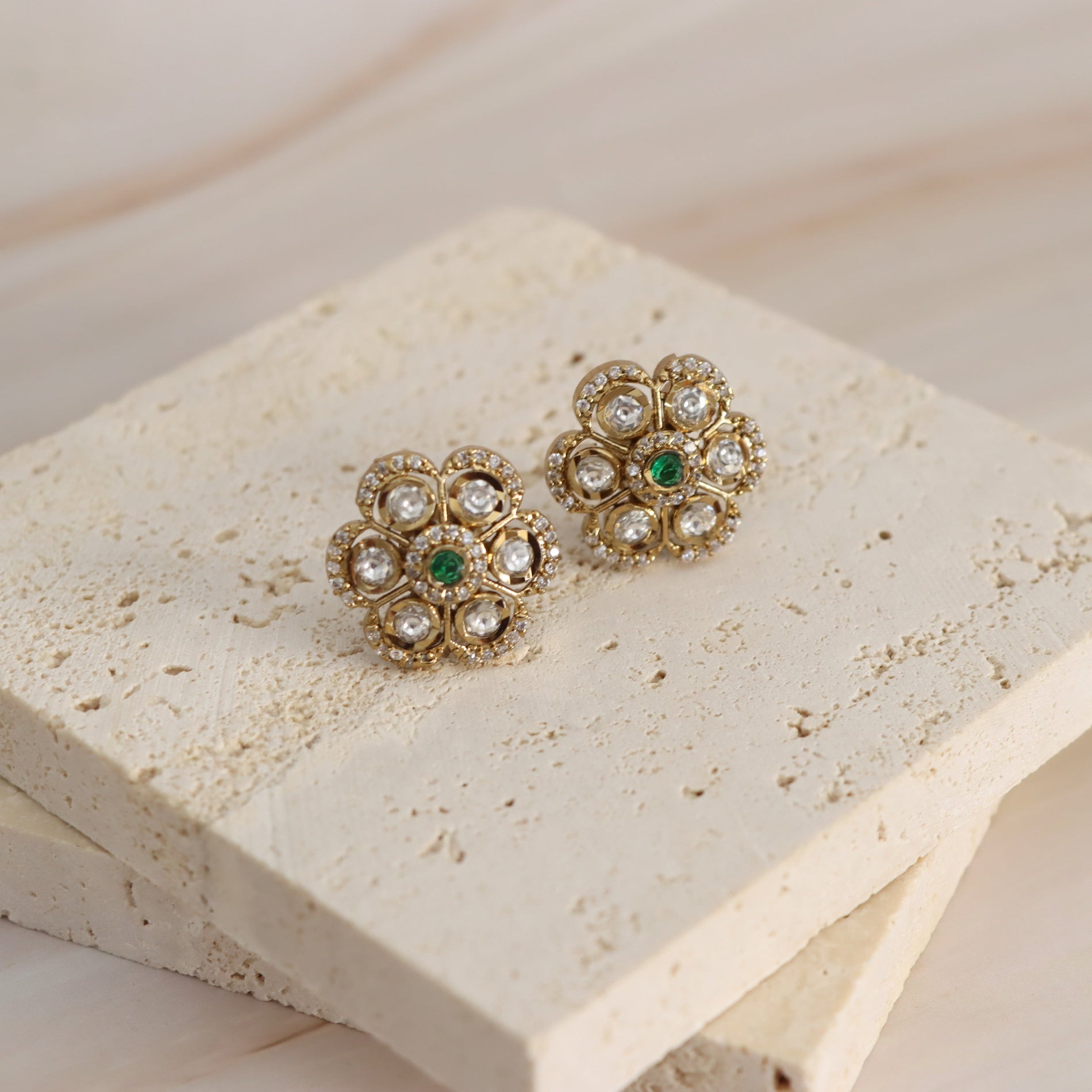 A pair of gold-toned floral stud earrings with clear rhinestones and small green center stones, displayed on a textured cream stone block against a light wood background.