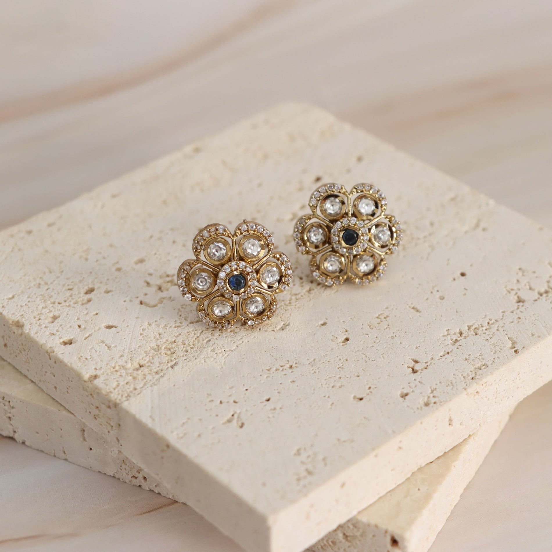 A pair of gold-toned floral stud earrings with clear rhinestones and dark blue center stones, displayed on stacked beige stone tiles.