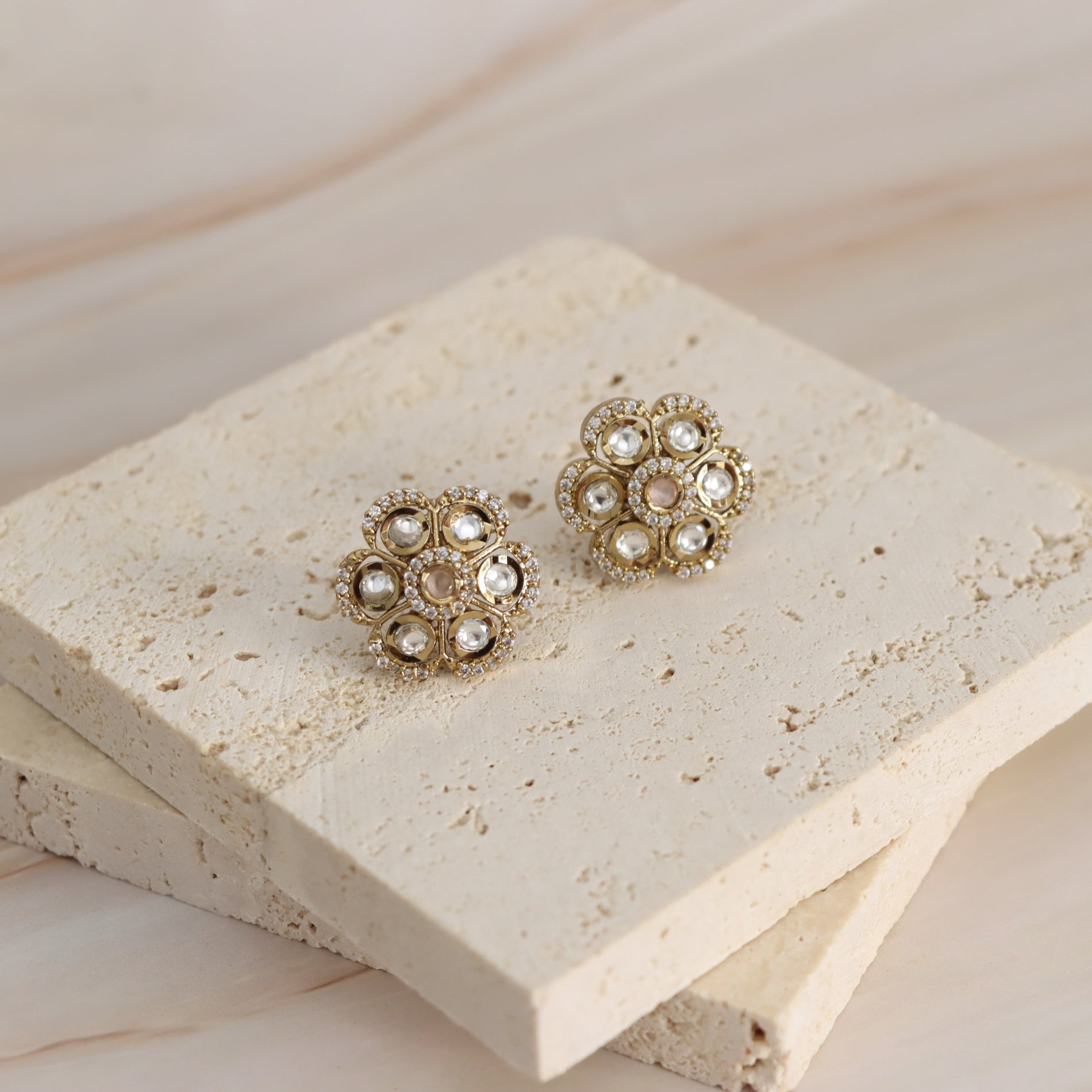 A pair of gold-toned floral stud earrings with clear rhinestones, displayed on stacked beige stone tiles against a light wood background.