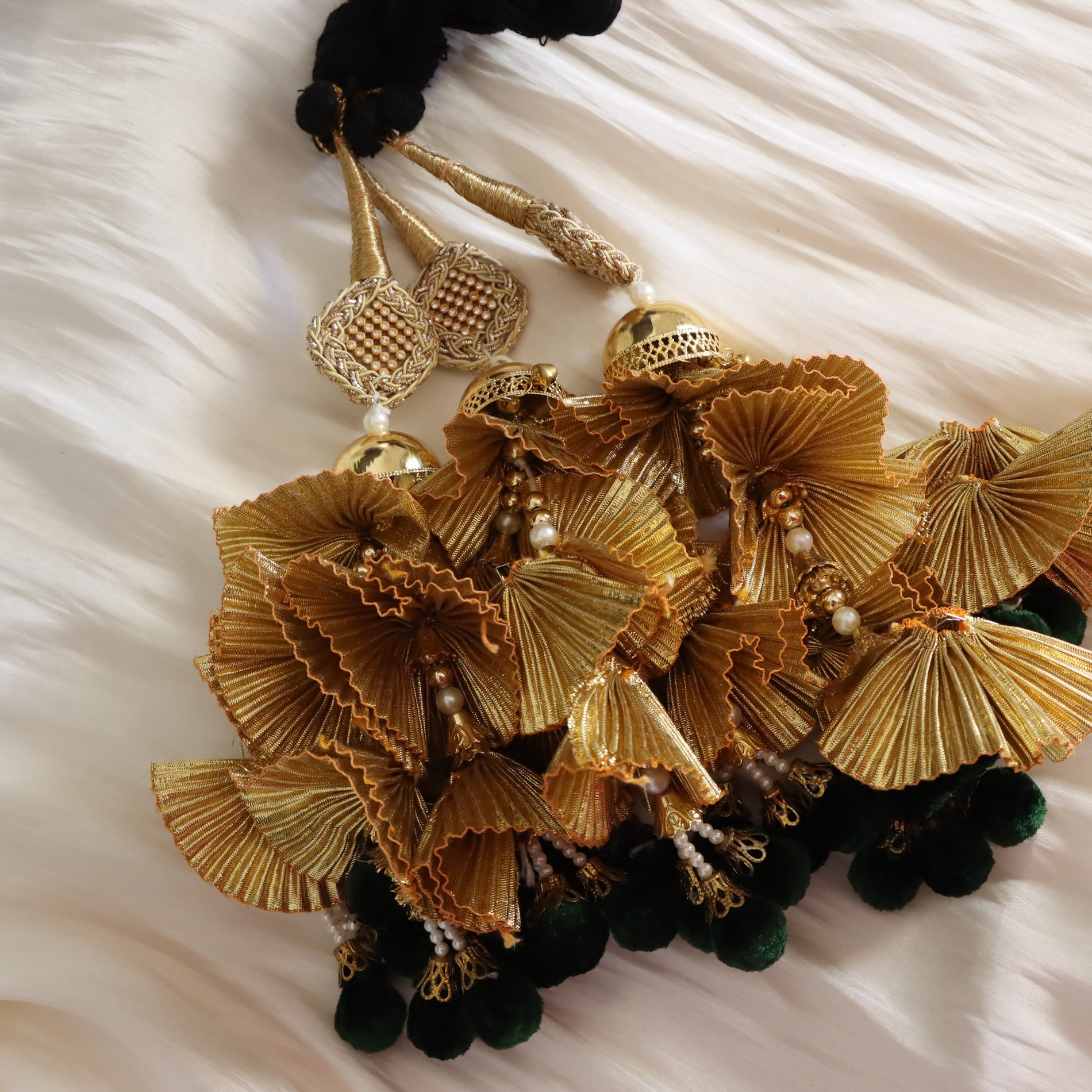 A close-up of ornate gold tassel jewelry with pleated fan-shaped elements, small pearl-like beads, and dark green pom-pom tassels, arranged on a soft white fabric background.