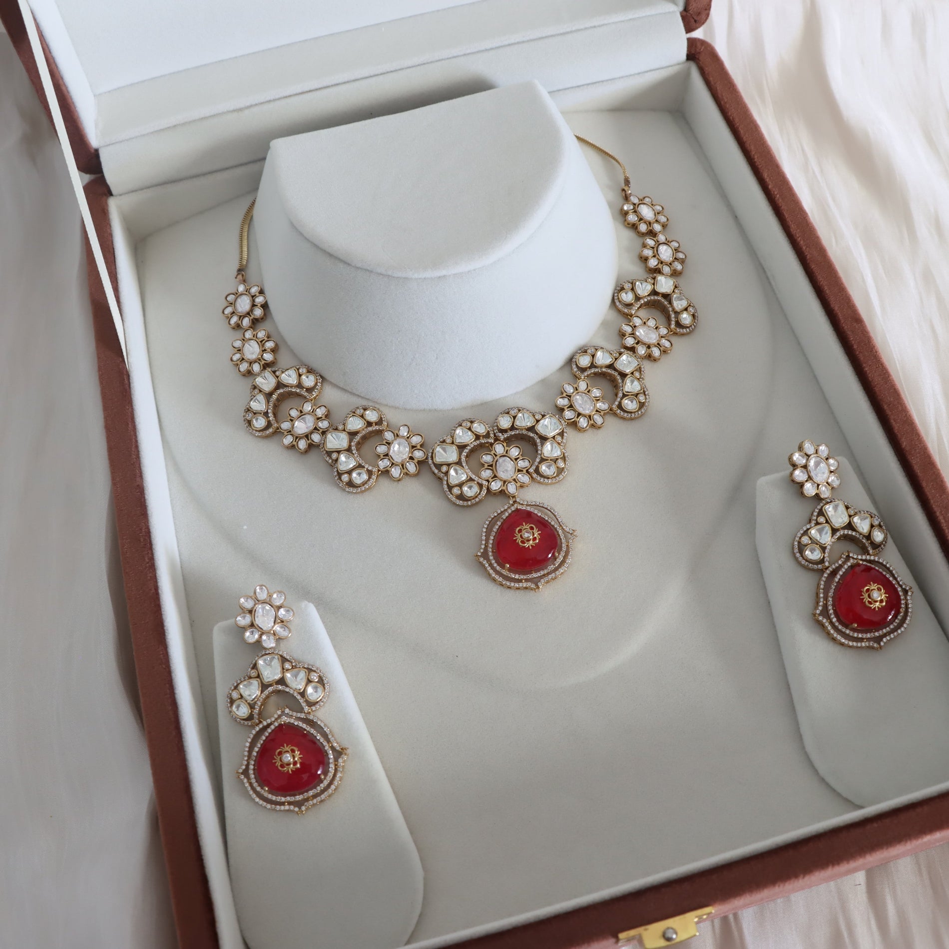 An ornate jewelry set displayed inside an open presentation box, featuring a gold-tone necklace with white stone floral motifs and a central red teardrop pendant, plus matching drop earrings on white display stands.
