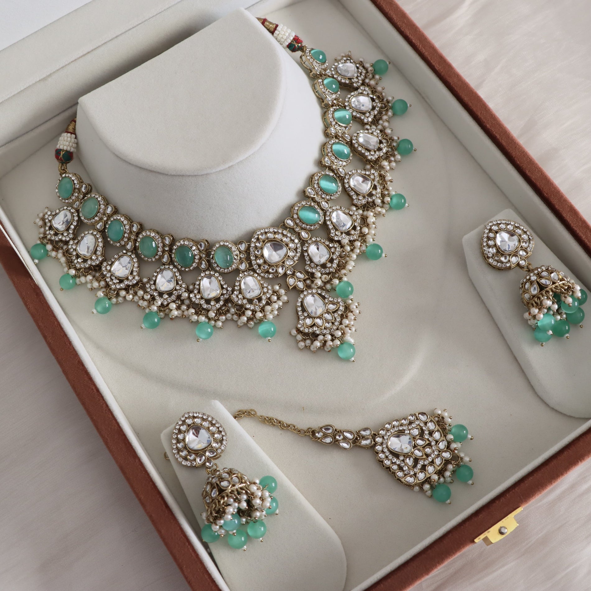 An ornate Indian traditional jewelry set displayed inside its brown presentation box. The set includes a gold-plated necklace with white pearls, clear crystals, and green stone accents, matching drop earrings, and a maang tikka, all arranged on cream-colored display stands.
