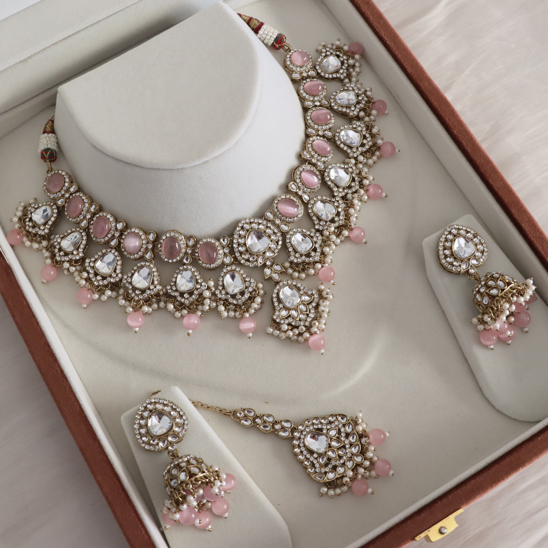 An ornate Indian jewelry set displayed inside its brown presentation box. The set includes a gold-plated necklace, matching earrings, and a maang tikka, all decorated with white pearls, pink beads, and clear rhinestones.