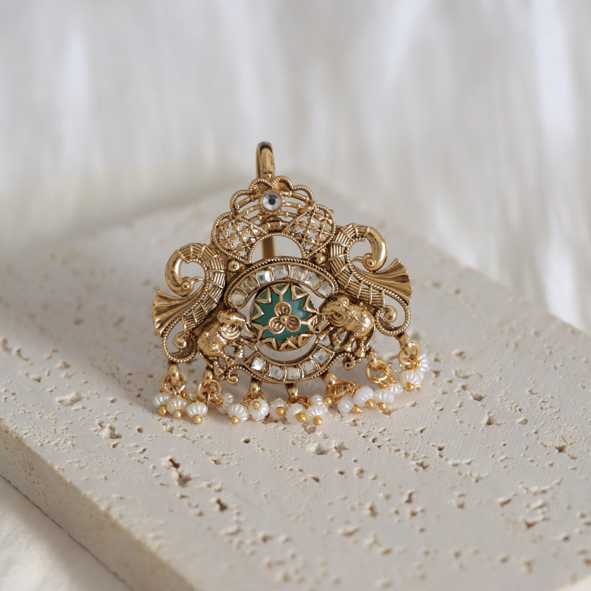 An ornate gold-toned pendant with intricate filigree detailing, a central star motif with teal and gold accents, and dangling pearl-like beads, displayed on a textured stone surface.