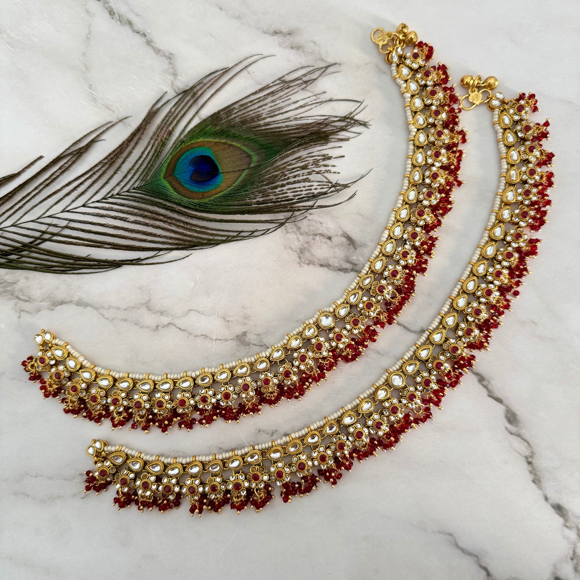 Gold and red anklet on a marble surface with a peacock feather