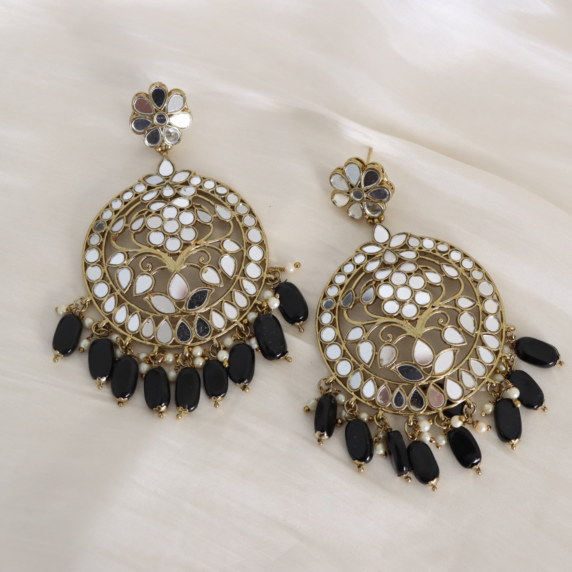 A pair of ornate gold-toned dangle earrings with white and black mirror work, pearl accents, and dangling black teardrop beads, displayed on a soft white fabric background.
