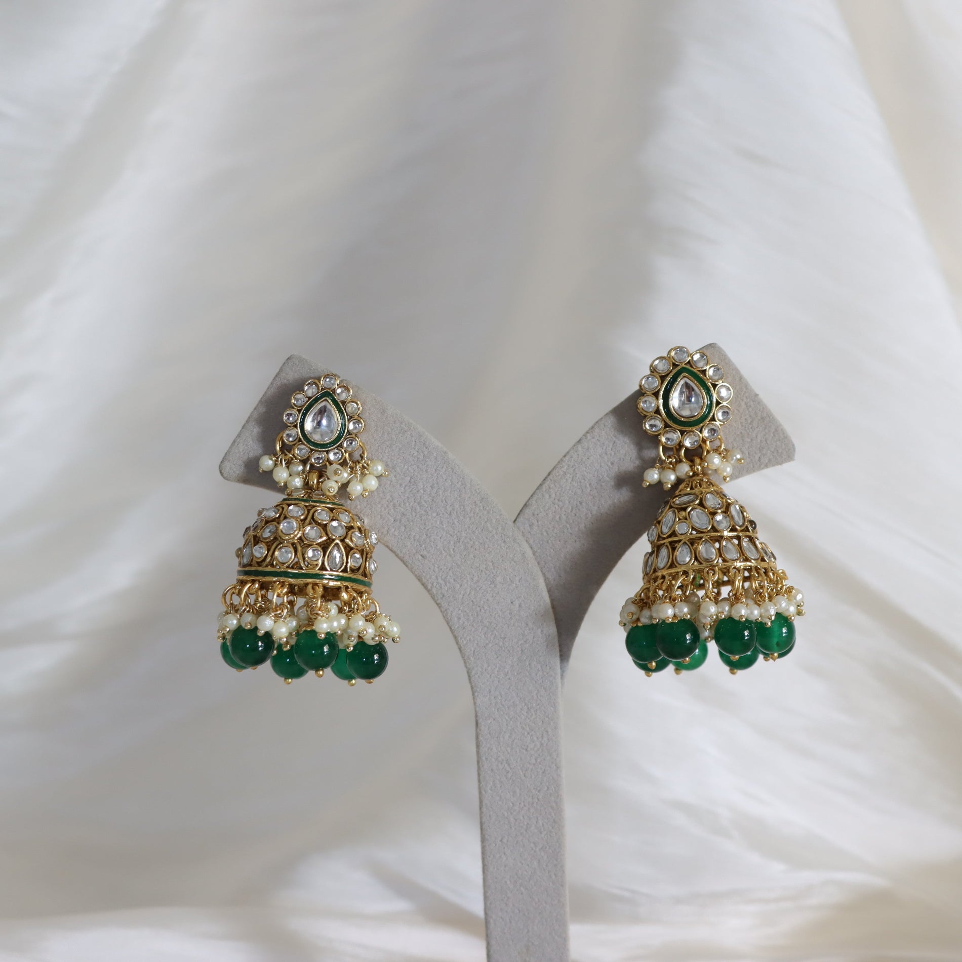 A pair of gold-plated Indian jhumka earrings with white faux pearls, green bead drops, and a central clear stone, displayed on a gray jewelry stand against a white fabric background.