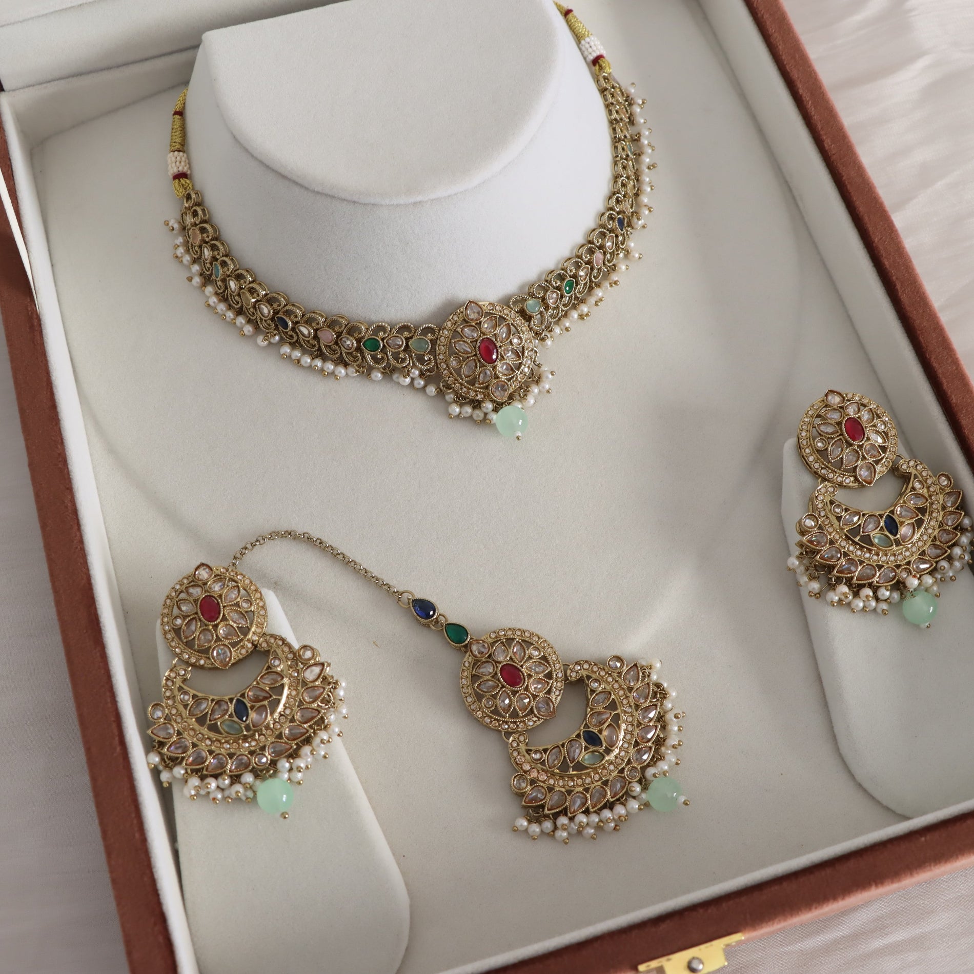 Gold-toned traditional Indian jewelry set displayed inside an open presentation box, including a choker necklace, matching earrings, and a maang tikka, decorated with white pearls, green stones, and red center stones.