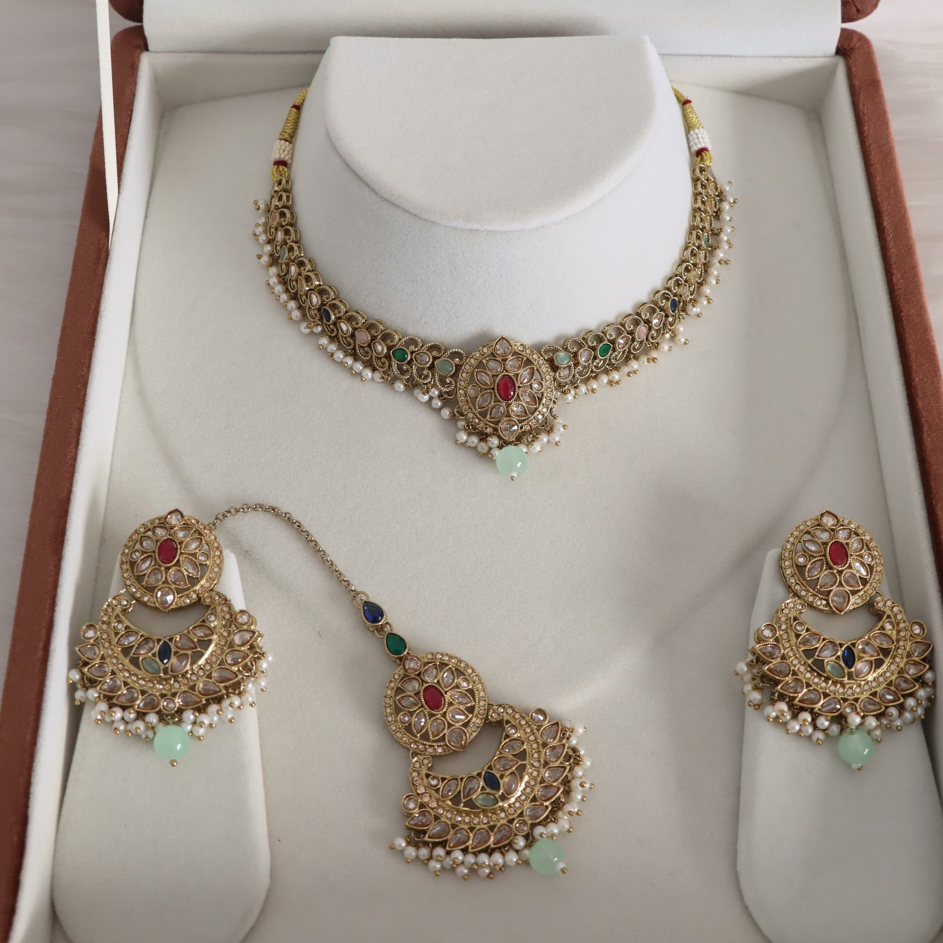 Gold-toned traditional Indian jewelry set displayed inside an open jewelry box, including a necklace on a white neck stand, a matching maang tikka, earrings, and a chain connecting them. The pieces feature gold filigree, white pearl-like beads, red and green stones, and light green bead drops.