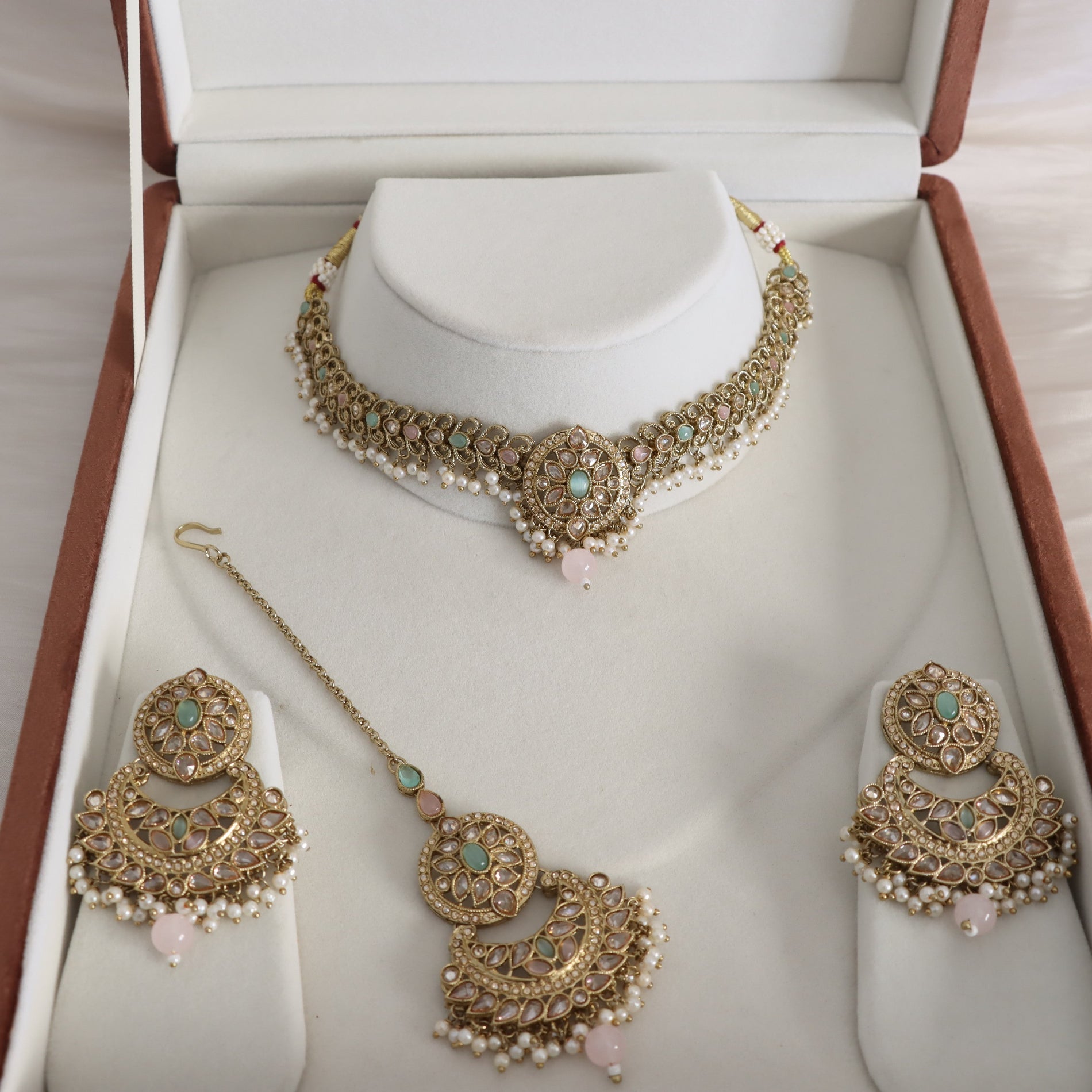 A traditional Indian gold-toned jewelry set displayed inside an open presentation box. The set includes a choker necklace with a central pendant, a matching maang tikka with a chain, and a pair of large chandelier earrings, all decorated with gold, white pearls, light green stones, and pink bead accents.