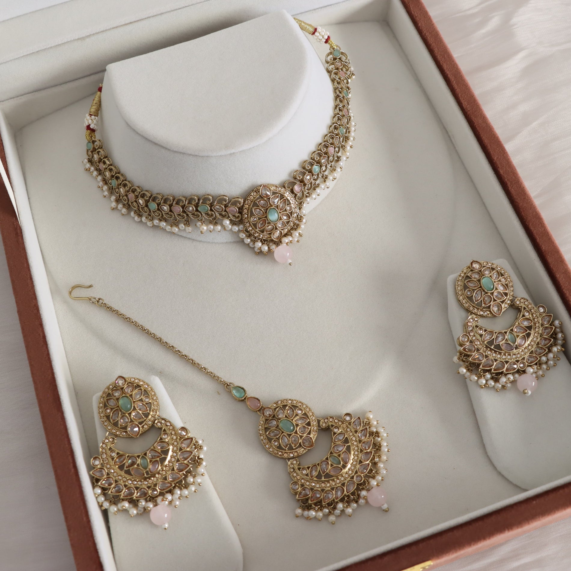 Gold-toned traditional Indian jewelry set including a choker necklace, matching earrings, and a maang tikka, displayed on a white jewelry box with pearl-like bead details and small pink stone accents.