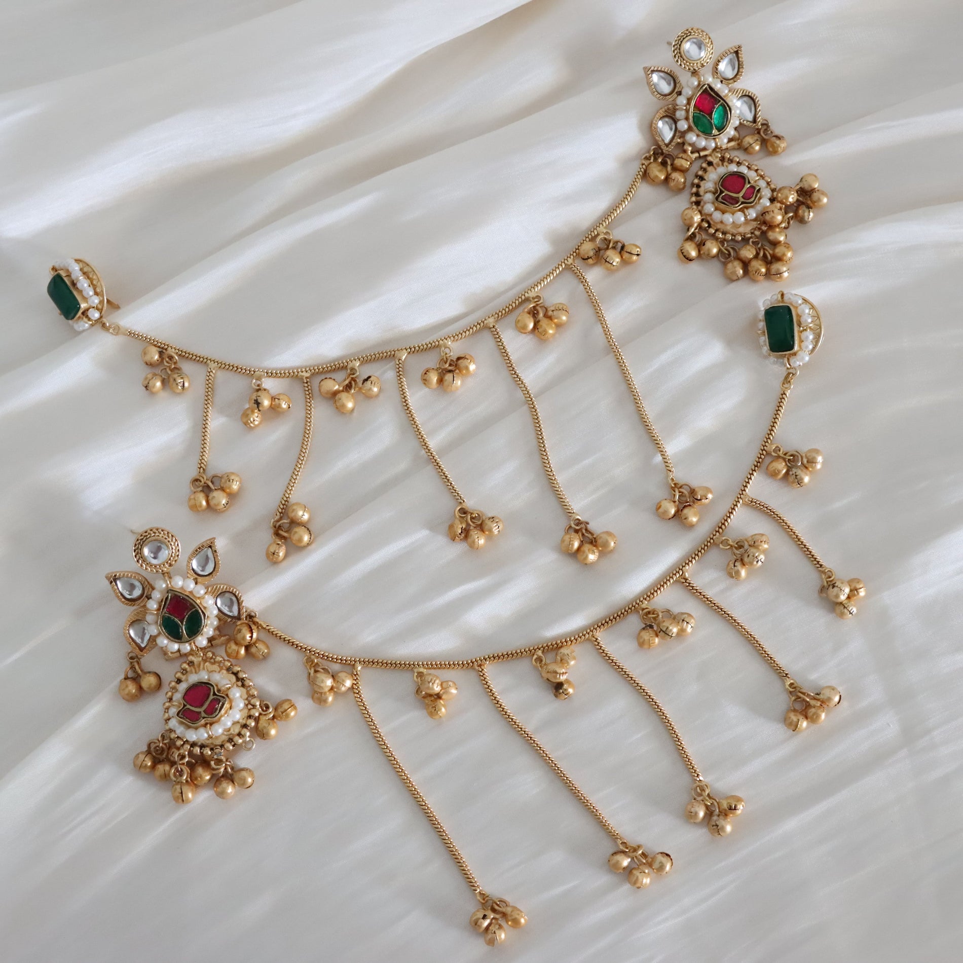 Gold-toned traditional Indian jewelry set featuring a chain with dangling beaded tassels and ornate floral motifs set with green stones, red stones, and clear stones, displayed on a white draped fabric background.
