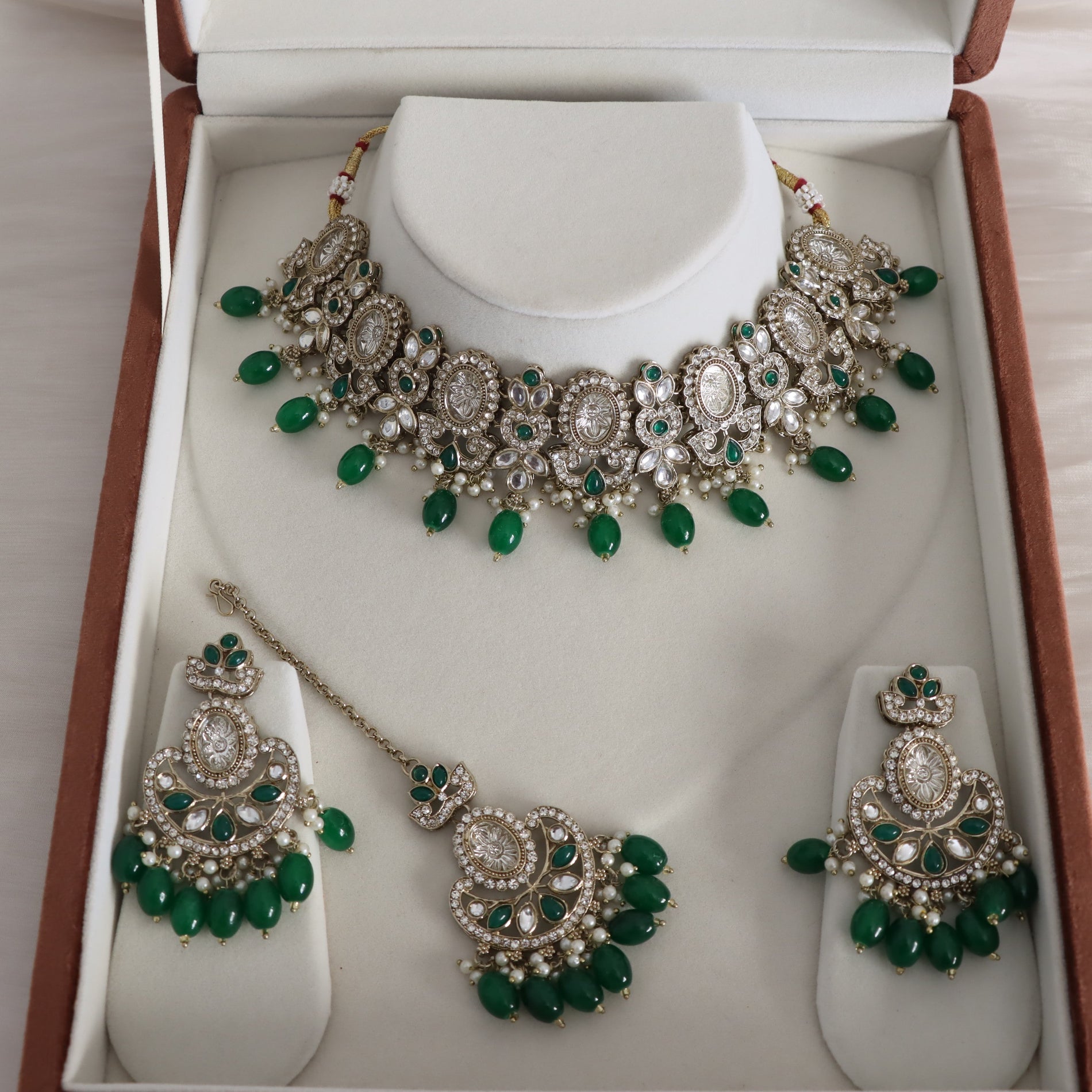 An ornate Indian traditional jewelry set displayed in its presentation box, featuring a silver-tone necklace, matching earrings, and a maang tikka with green gemstone accents.