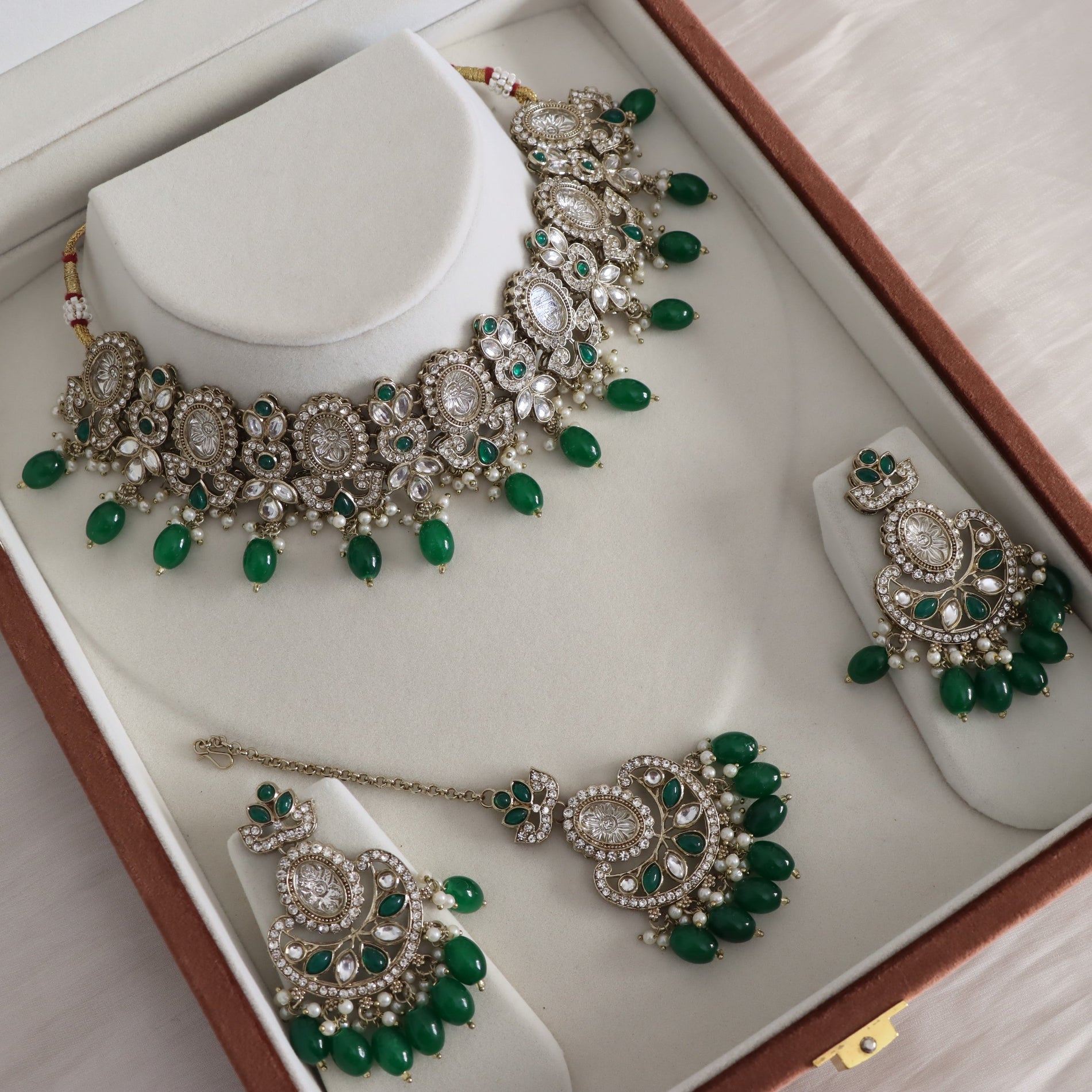 An ornate Indian ethnic jewelry set displayed inside a brown presentation box. The set includes a silver-toned necklace, matching earrings, and a maang tikka, all decorated with white faux pearls, clear crystals, and vibrant green bead accents.