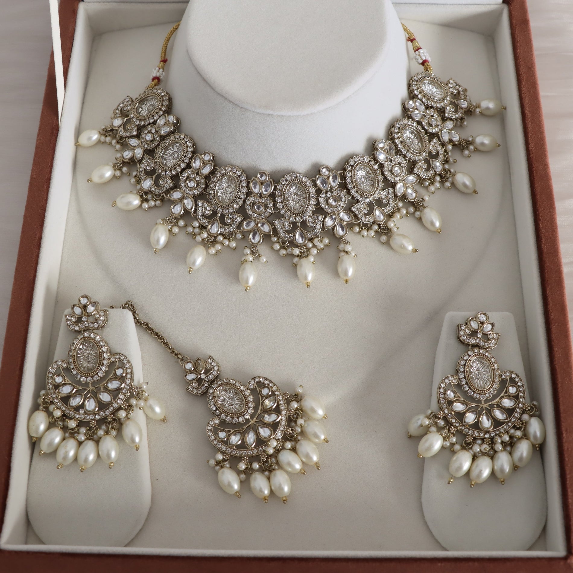 A traditional Indian jewelry set displayed in its presentation box, featuring a choker necklace, a matching pendant, and earrings. The jewelry is embellished with silver-toned metal, clear crystals, and white faux pearls.