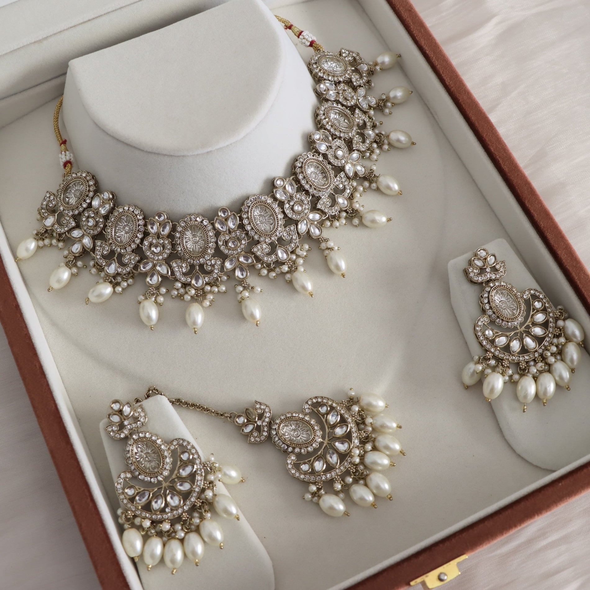 An ornate Indian ethnic jewelry set displayed inside its brown presentation box. The set includes a silver-toned statement necklace with white pearls, matching drop earrings, and a choker, all arranged on white display stands.