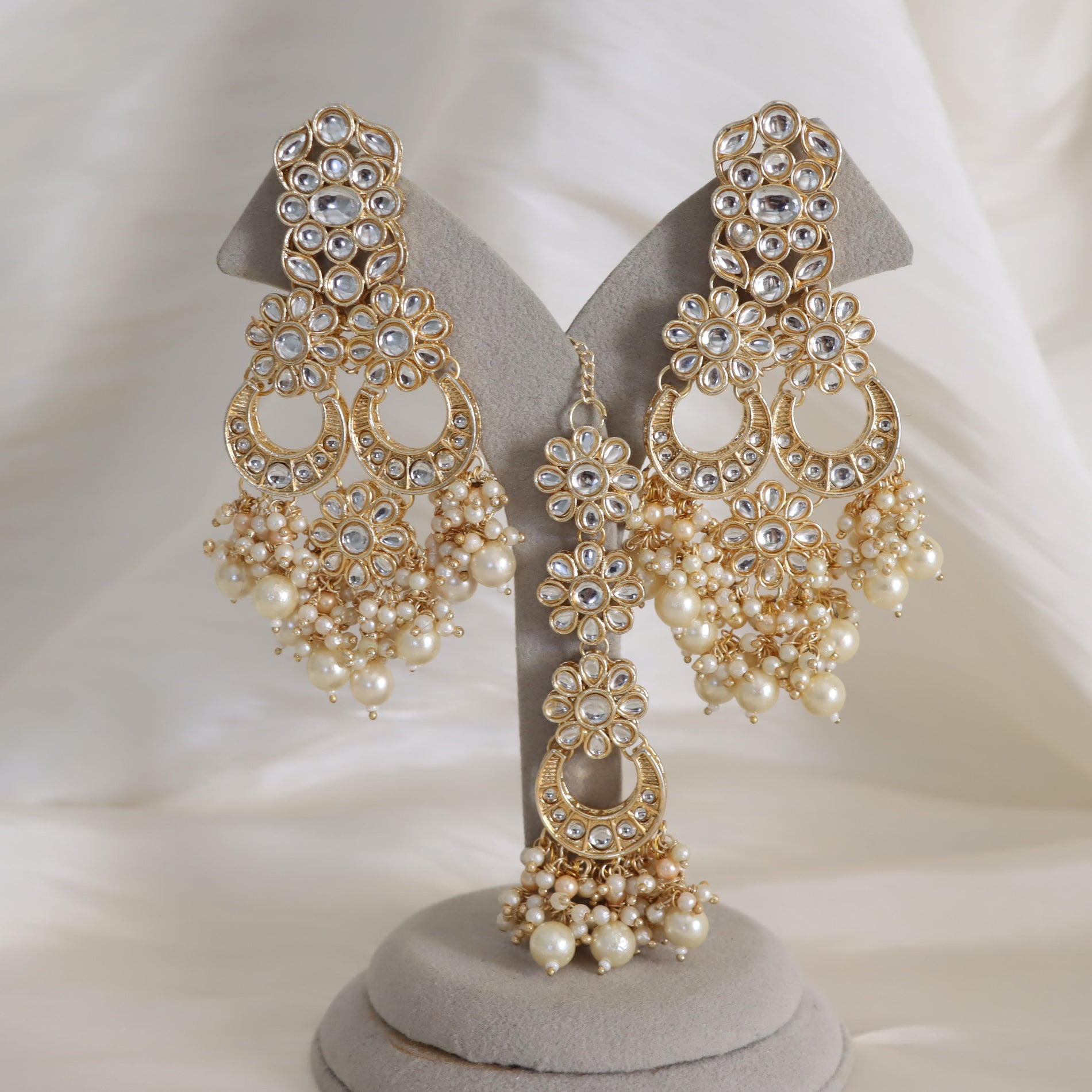 A pair of ornate gold-toned chandelier earrings with clear rhinestones and cream pearl drops, displayed on a gray jewelry stand against a soft white draped fabric background.