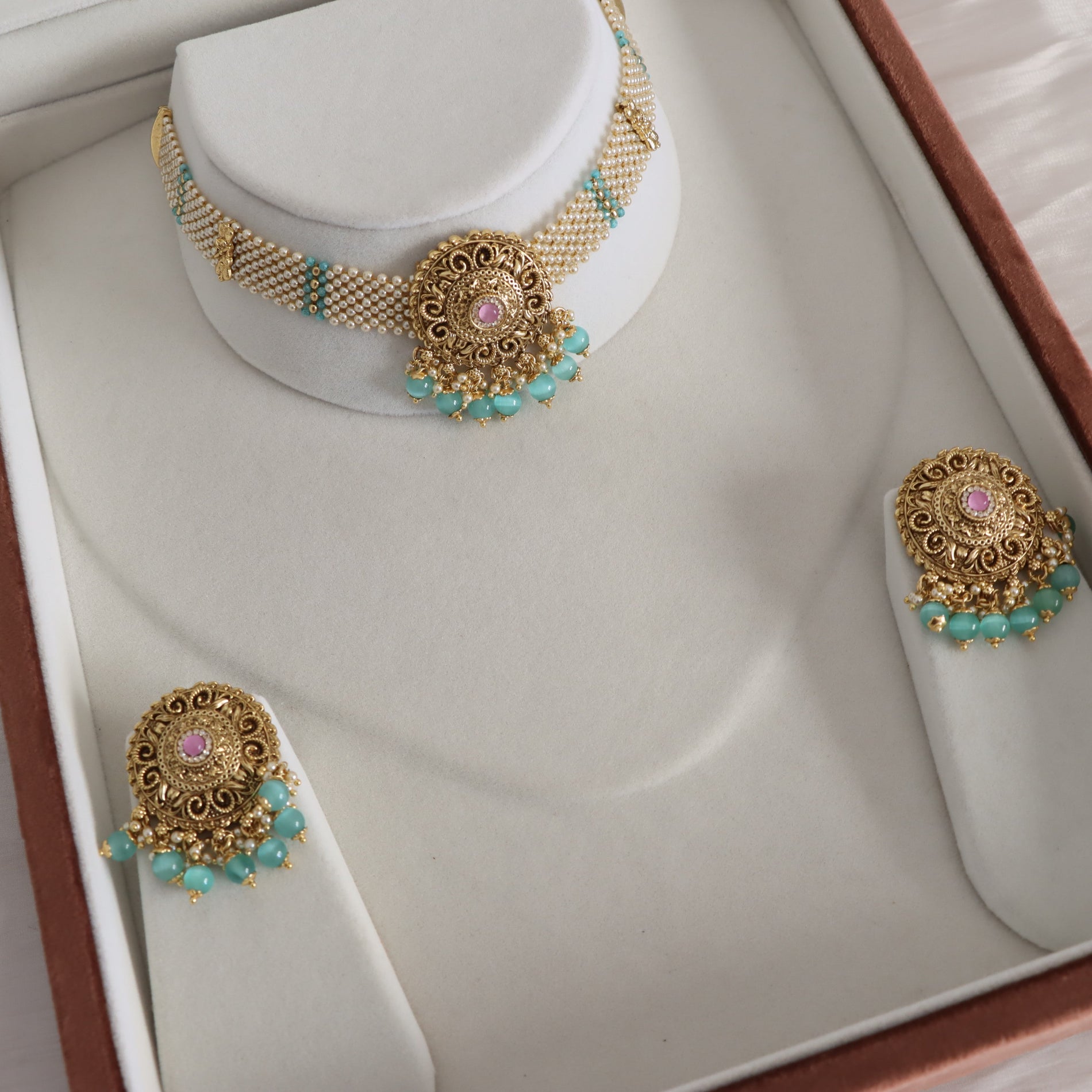 Gold-toned traditional jewelry set displayed on a cream jewelry box, including a choker necklace, earrings, and a ring, featuring turquoise bead accents and pink stone centers.