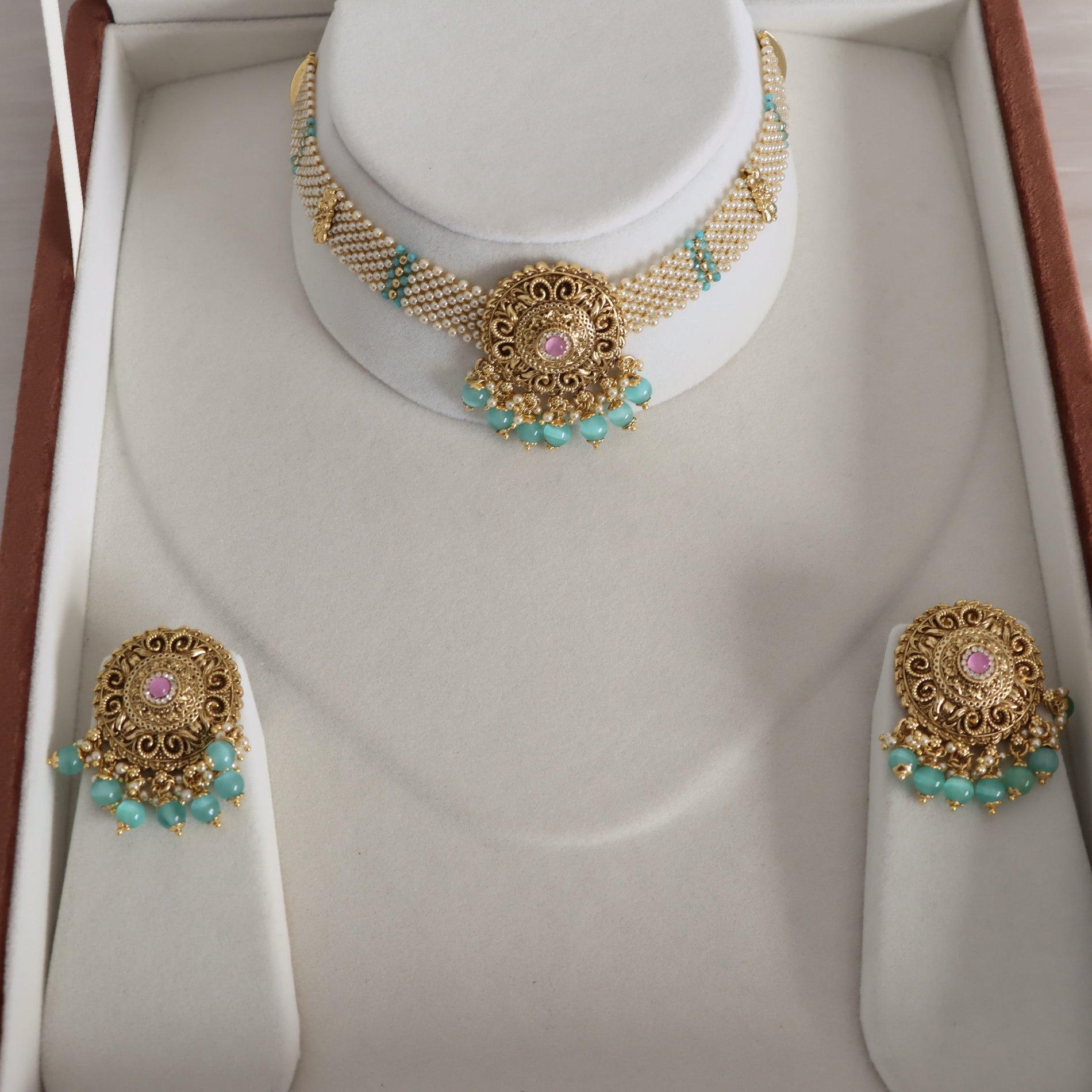 Gold-toned traditional jewelry set displayed in a white presentation box, including a choker necklace with a central floral motif and matching earrings, decorated with turquoise beads and pink stone accents.