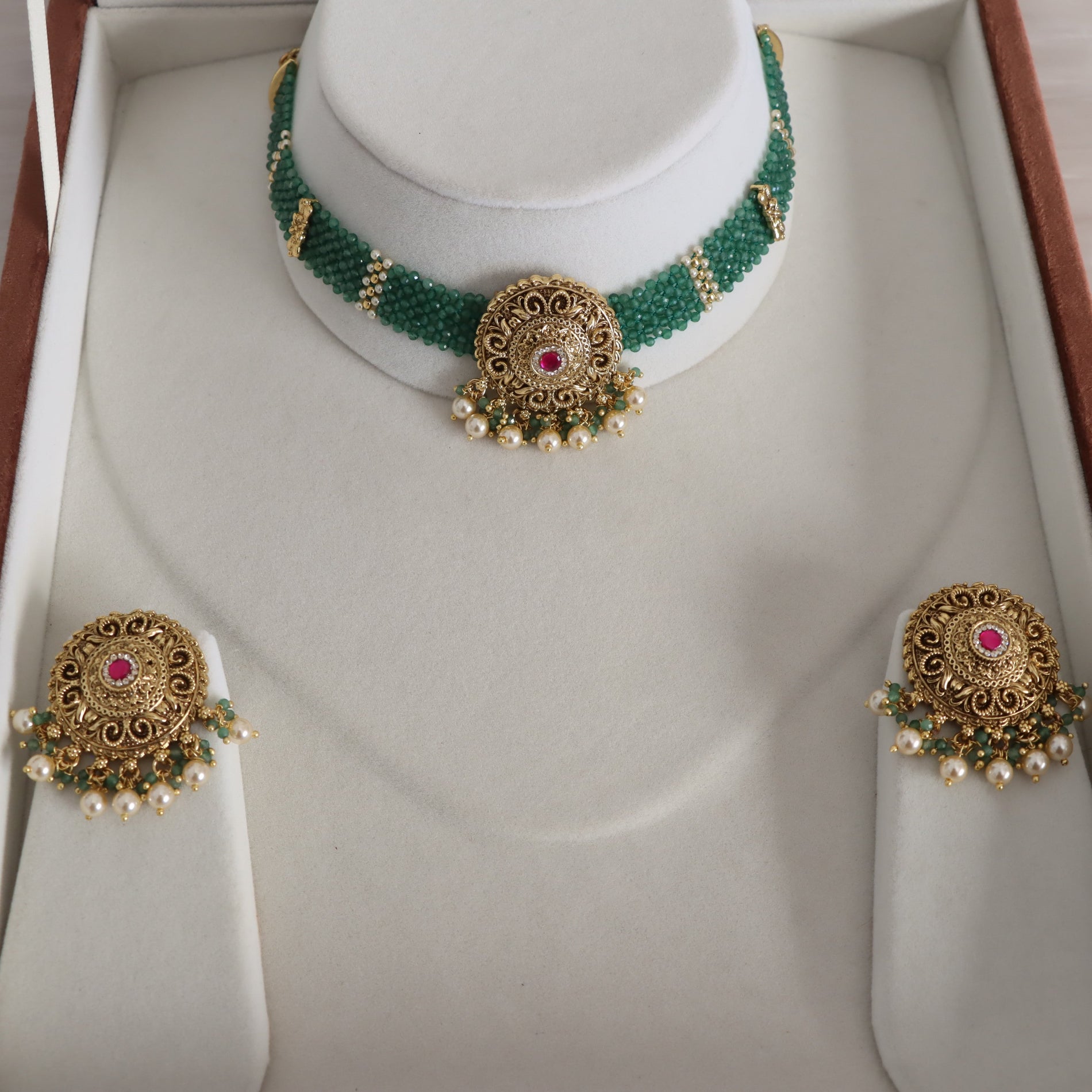 A traditional Indian jewelry set displayed on a white jewelry box, featuring a beaded green and gold choker necklace with a central gold filigree medallion, matching gold earrings, and pearl accents.