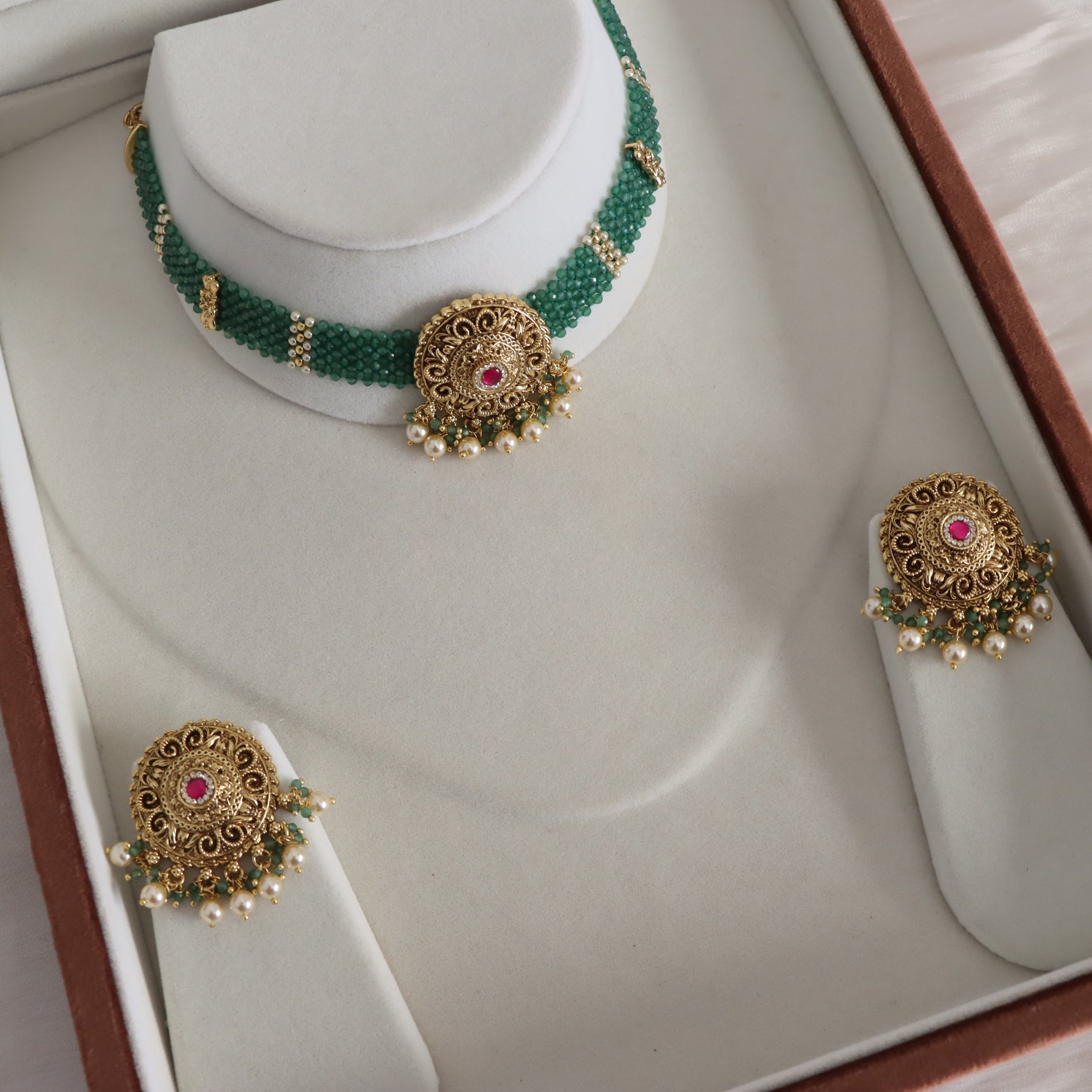 A traditional Indian jewelry set displayed in a white jewelry box, featuring a green and gold beaded necklace with a central gold filigree pendant with a red stone and pearl accents, along with matching earrings.